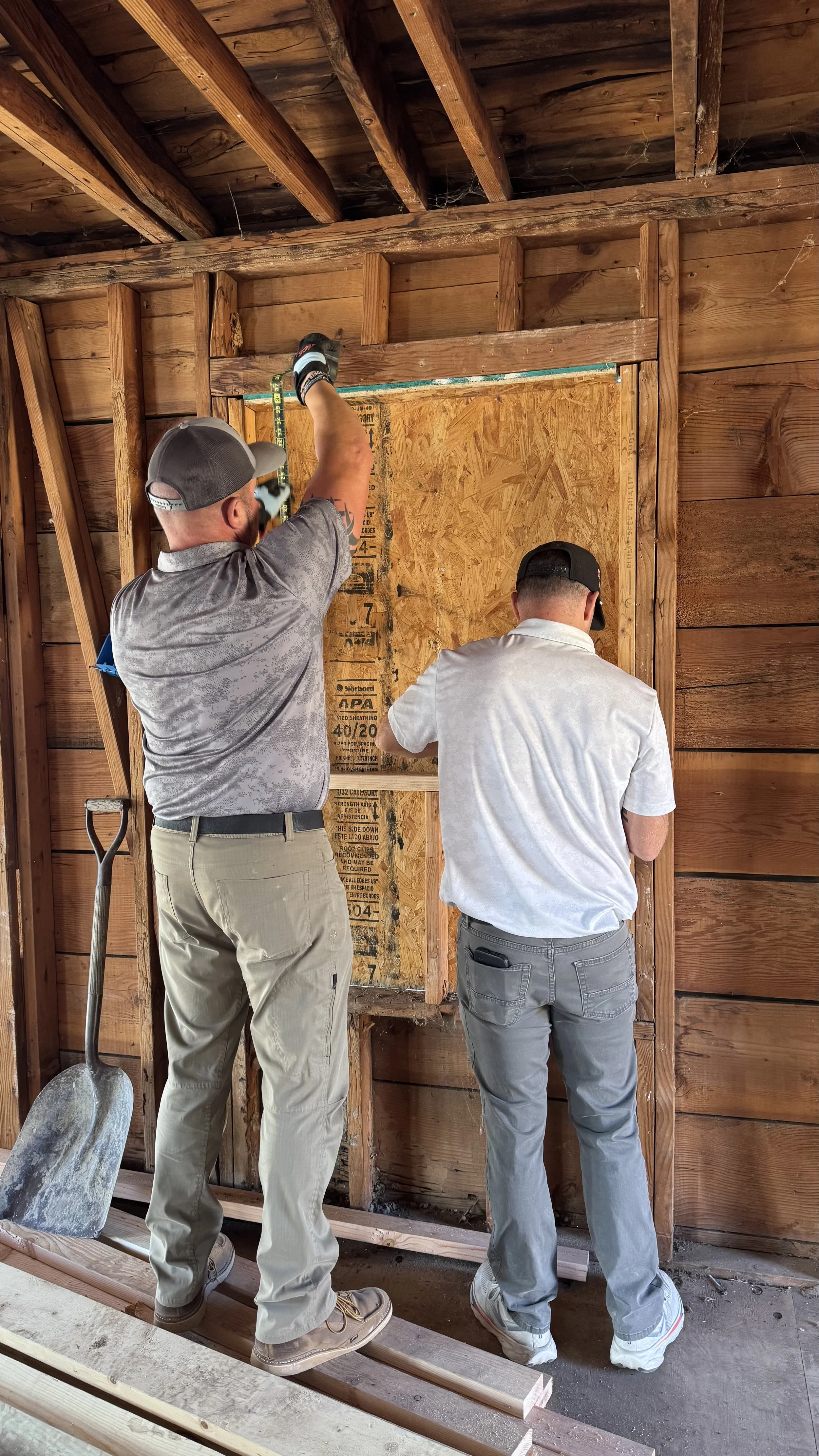 Two men are installing a wooden wall frame inside a building, with tools and construction materials around them.