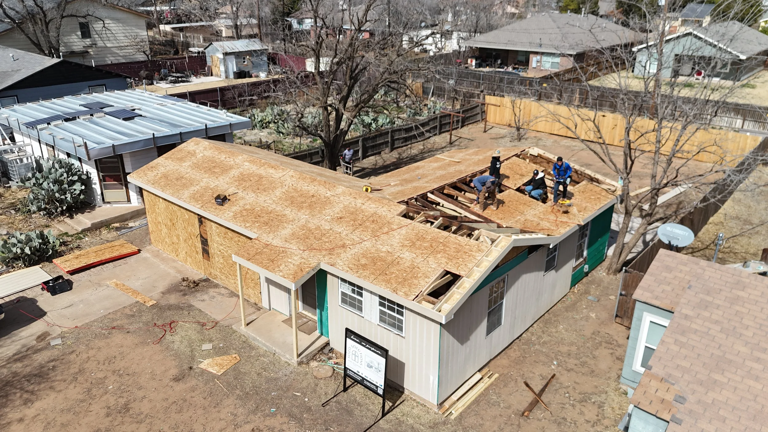 A house under construction with workers installing the roof, some sections covered with plywood sheathing, in a residential neighborhood.
