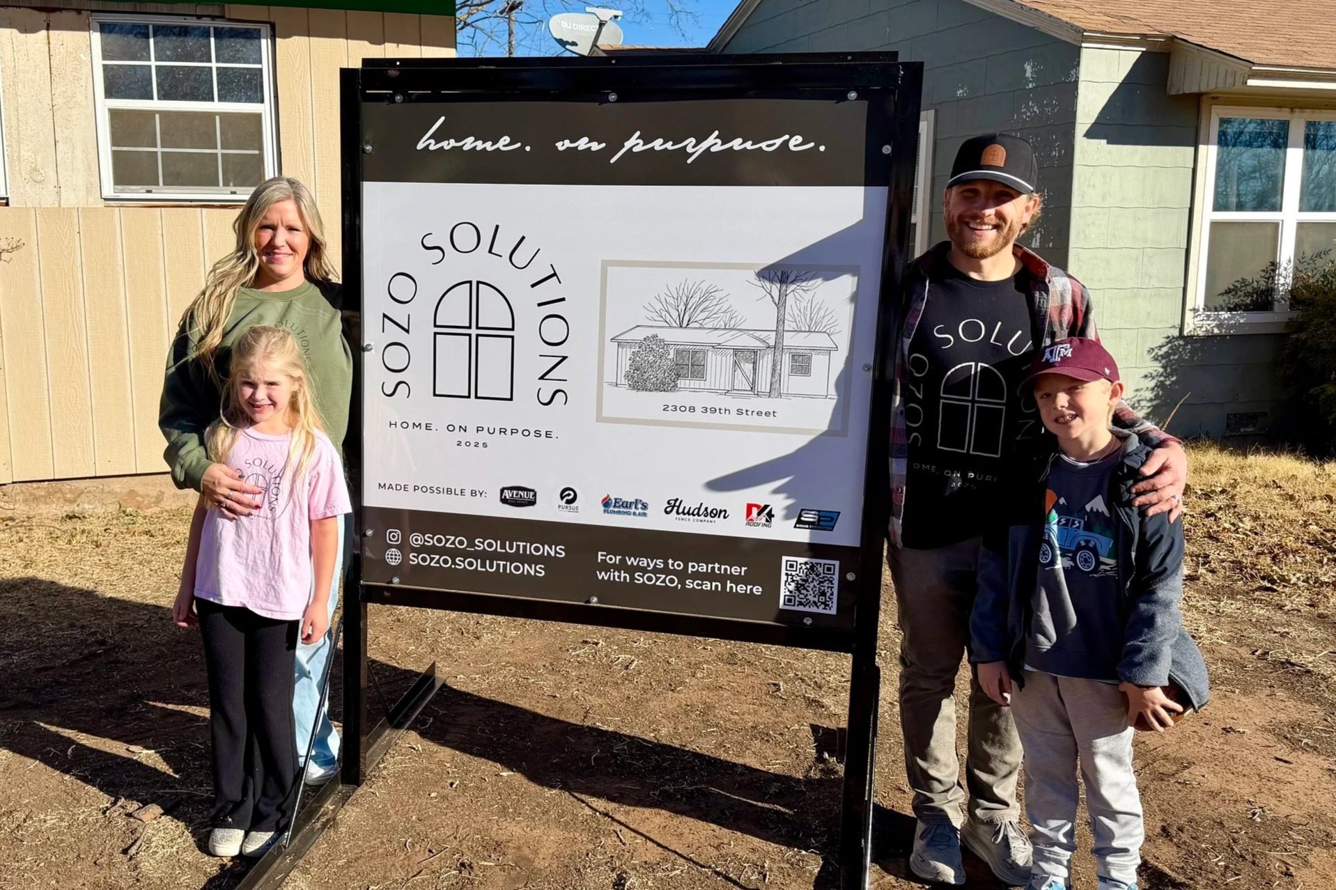 A family of four standing outdoors next to a large sign for SOZO Solutions. The sign features a logo with a window and include details about their home solutions business, with a drawing of a house and trees, their address, and social media information. The family includes a woman, two children (a girl and a boy), and a man, all smiling and standing on a dirt ground in front of a house with windows and lawn.