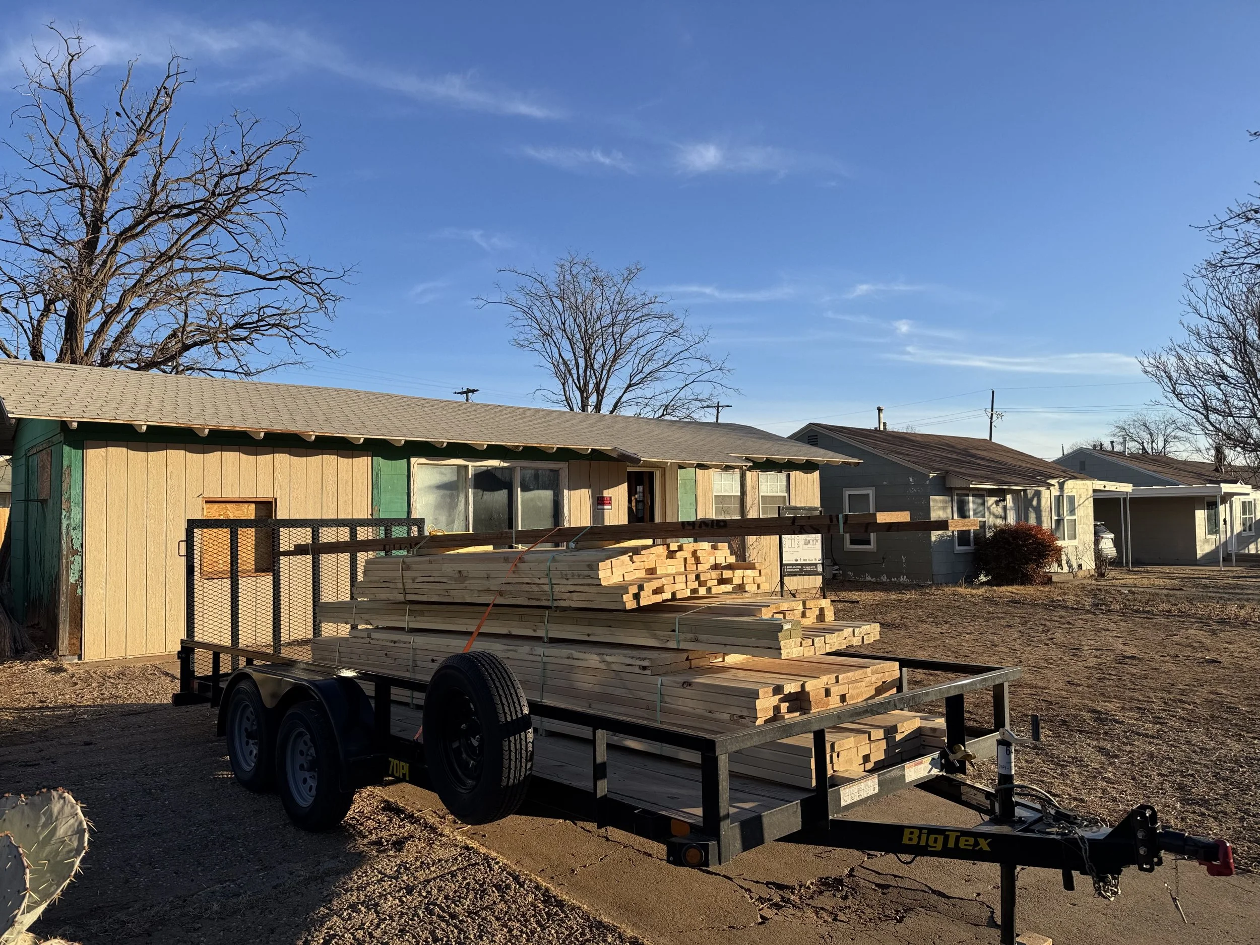 Trailer loaded with stacks of lumber parked in front of a row of neighborhood houses under a clear blue sky.