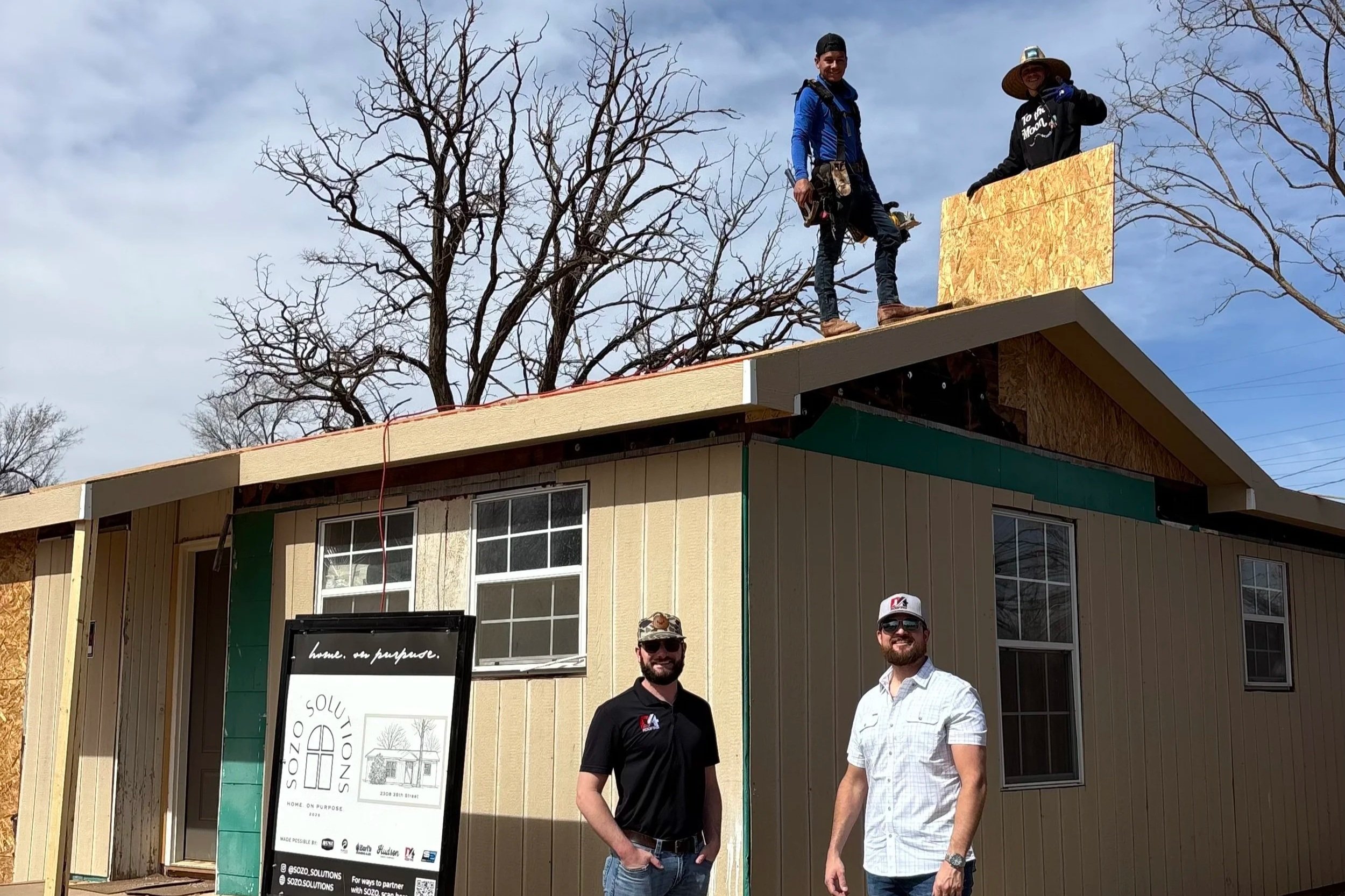 Three people on the roof of a house under construction, with two on the ground standing in front of the house. The house is partially built with windows and siding, and construction tools and materials are visible. The sky is cloudy, and there are leafless trees in the background.