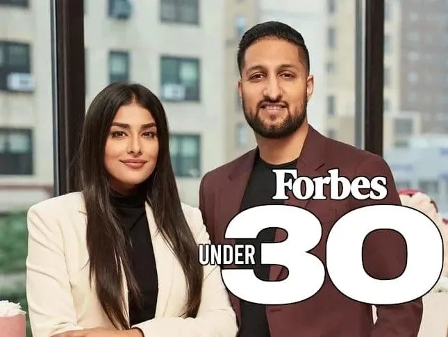 Two people standing indoors, with a "Forbes Under 30" logo displayed.