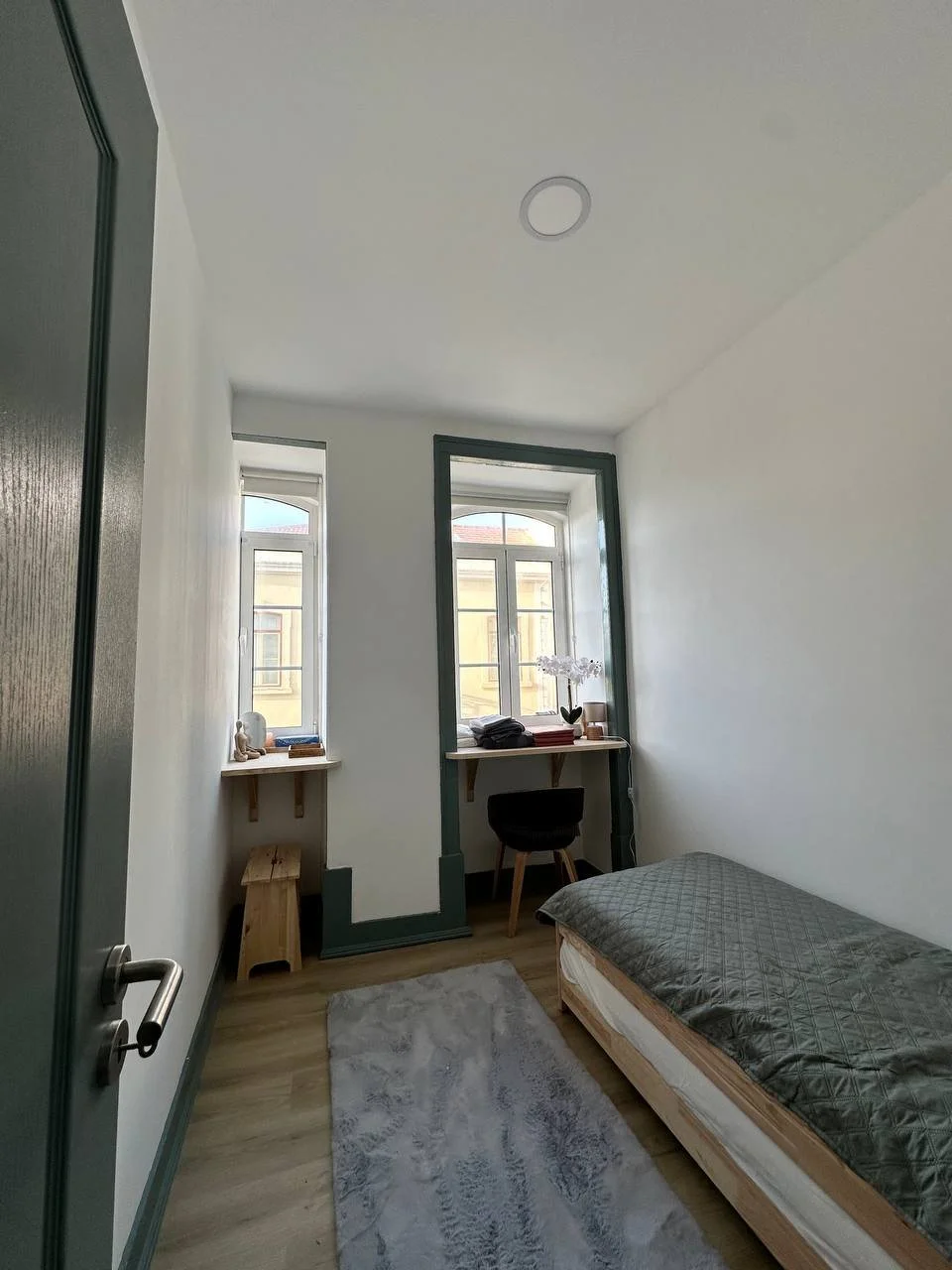 Small bedroom with single bed, desk, and chair, featuring two windows, green accents, and a light gray rug.