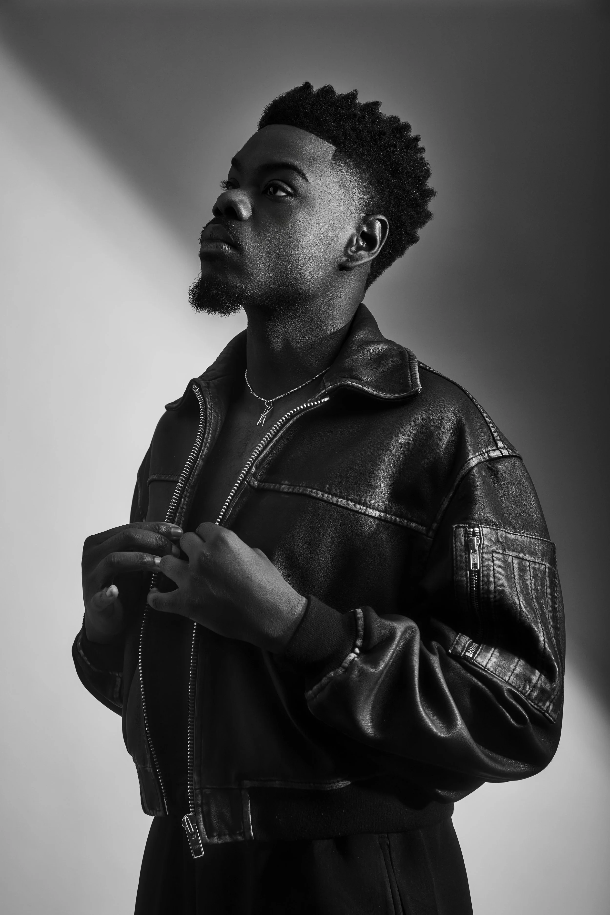 A black and white portrait of a young man with short, curly hair and a beard, wearing a leather jacket and a necklace, looking slightly upward with a serious expression.