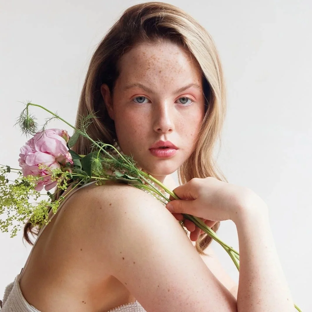 A young woman with red hair and freckles holding a bouquet of pink and green flowers, looking directly at the camera against a plain white background.