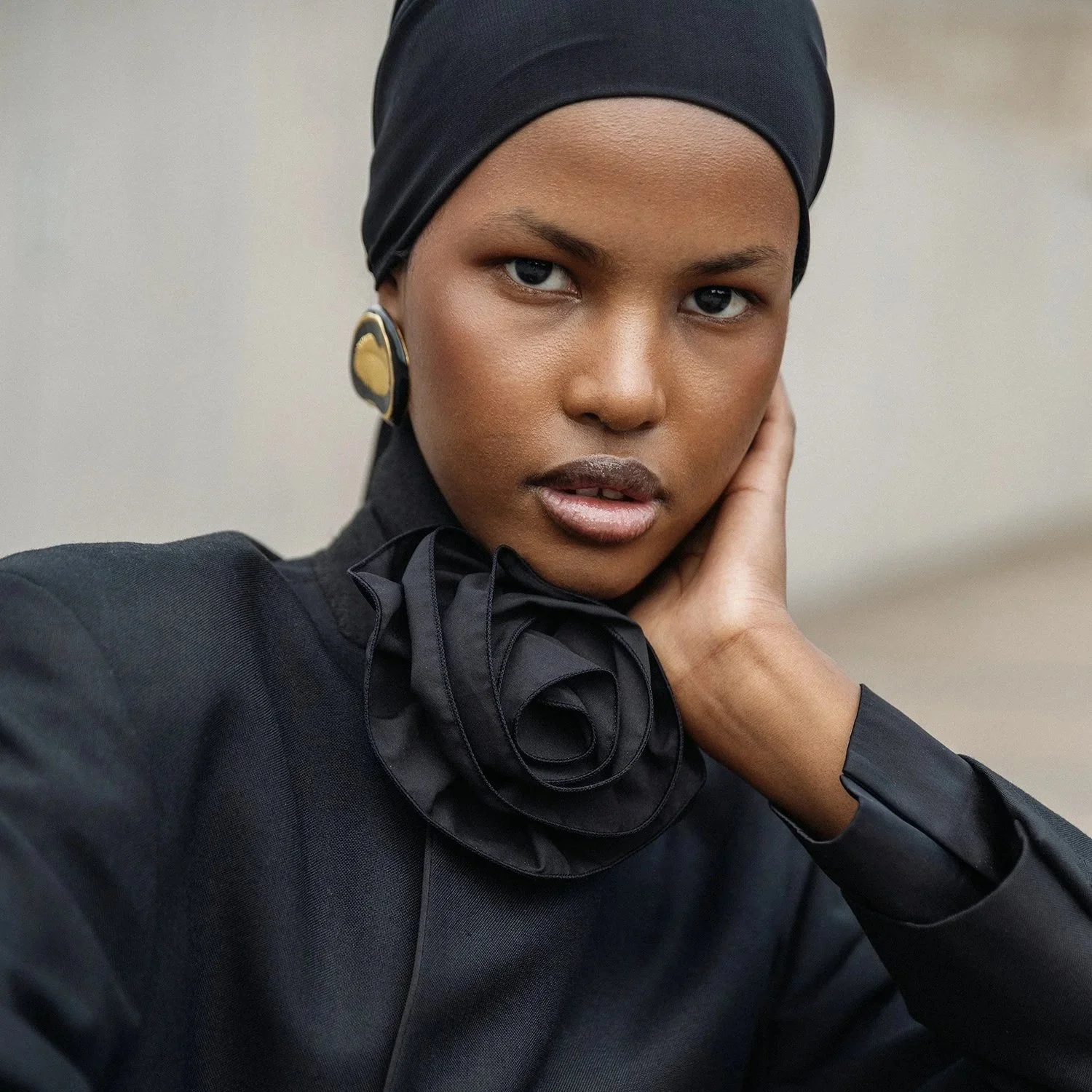 A woman wearing a black headscarf, black outfit with a ruffled neck detail, and large gold and black earrings, resting her hand on her neck and looking into the camera.