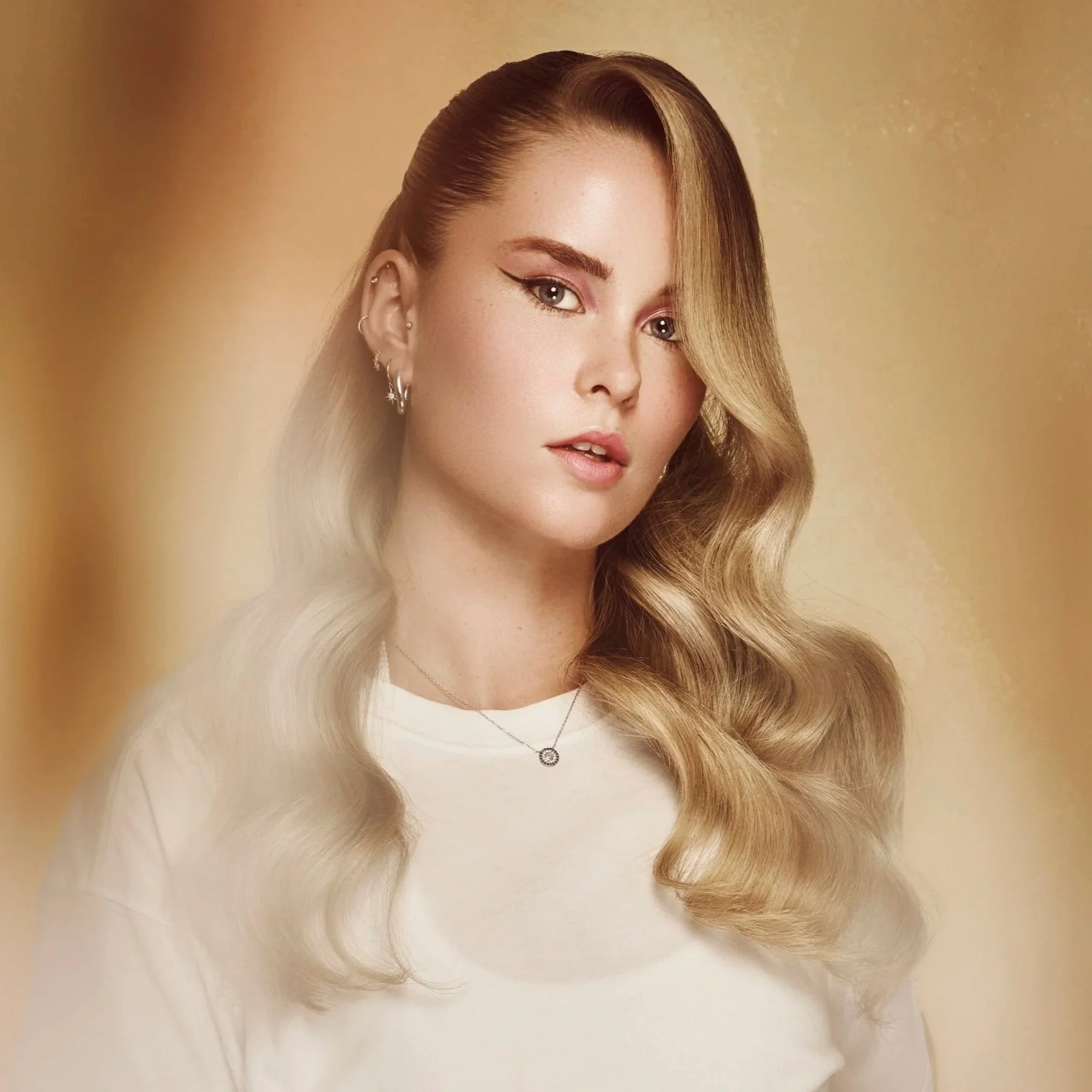 A young woman with long, wavy blonde hair wearing a white top, multiple earrings, and a necklace, against a warm beige background.
