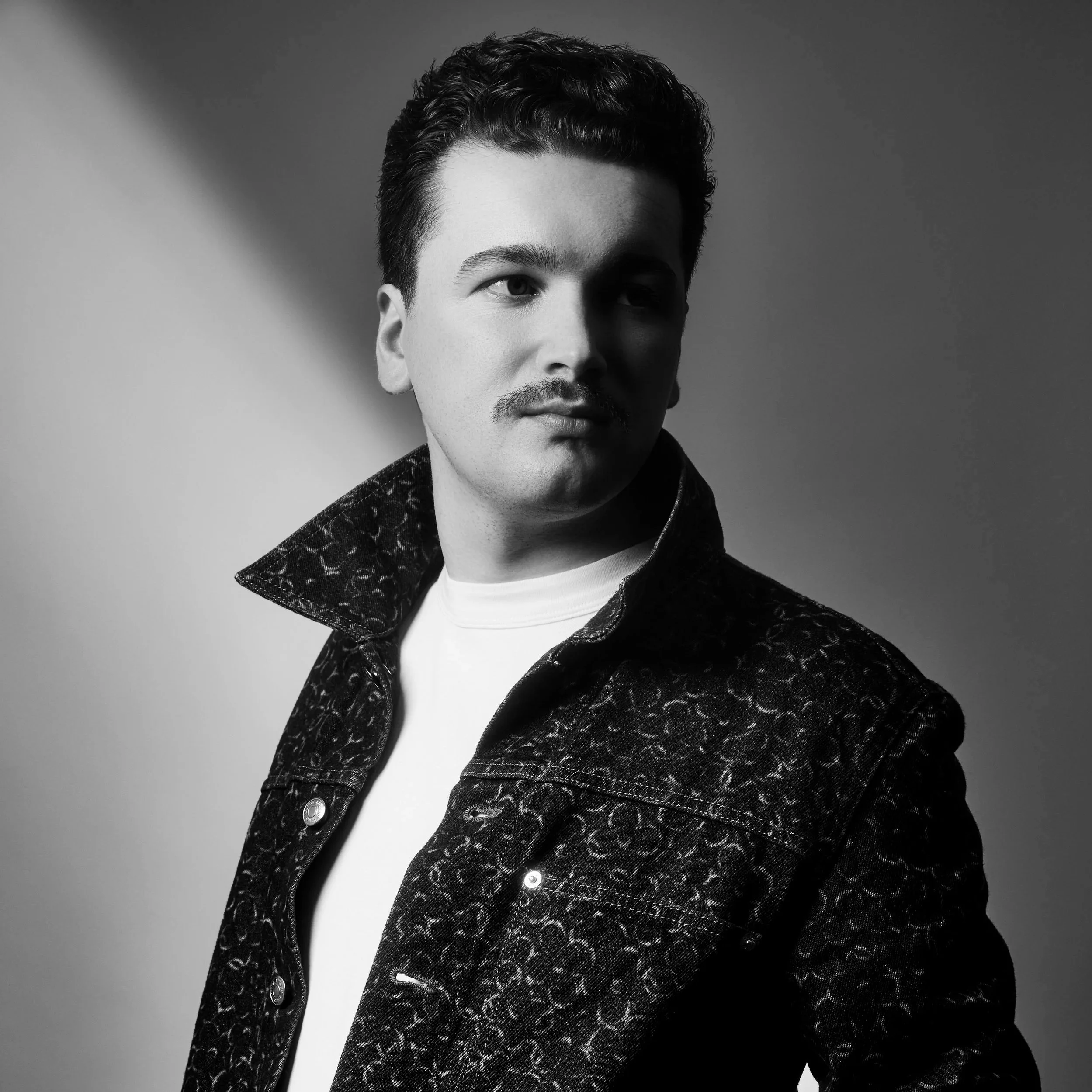 Black and white portrait of a man with dark hair, mustache, wearing a patterned dark jacket and a white shirt, looking to the side.