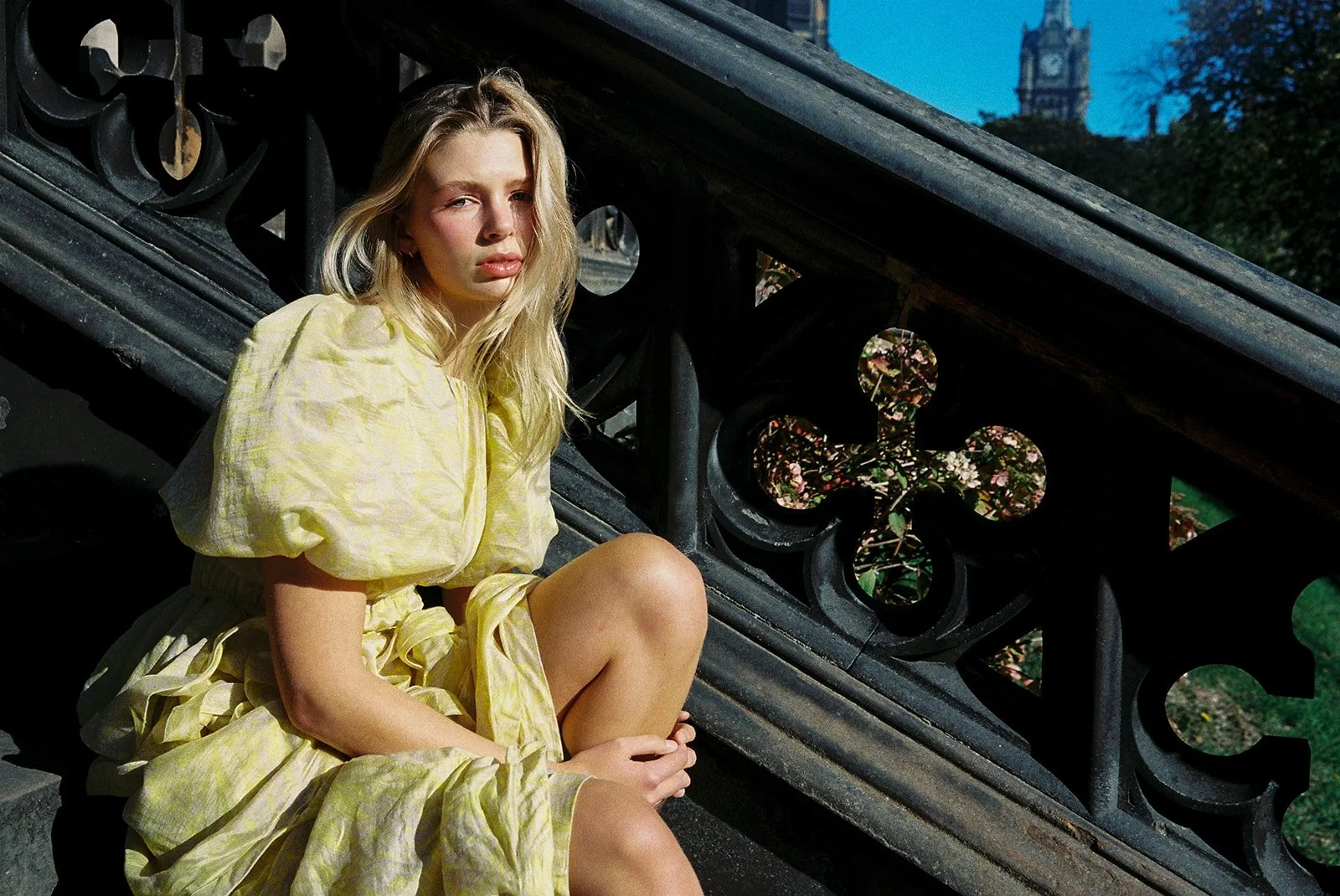 A young woman with blonde hair is sitting on stairs next to a black railing with decorative cutouts. She is wearing a yellow dress and is holding her leg with her hands. There is a building with a clock tower and a tree in the background on a clear e