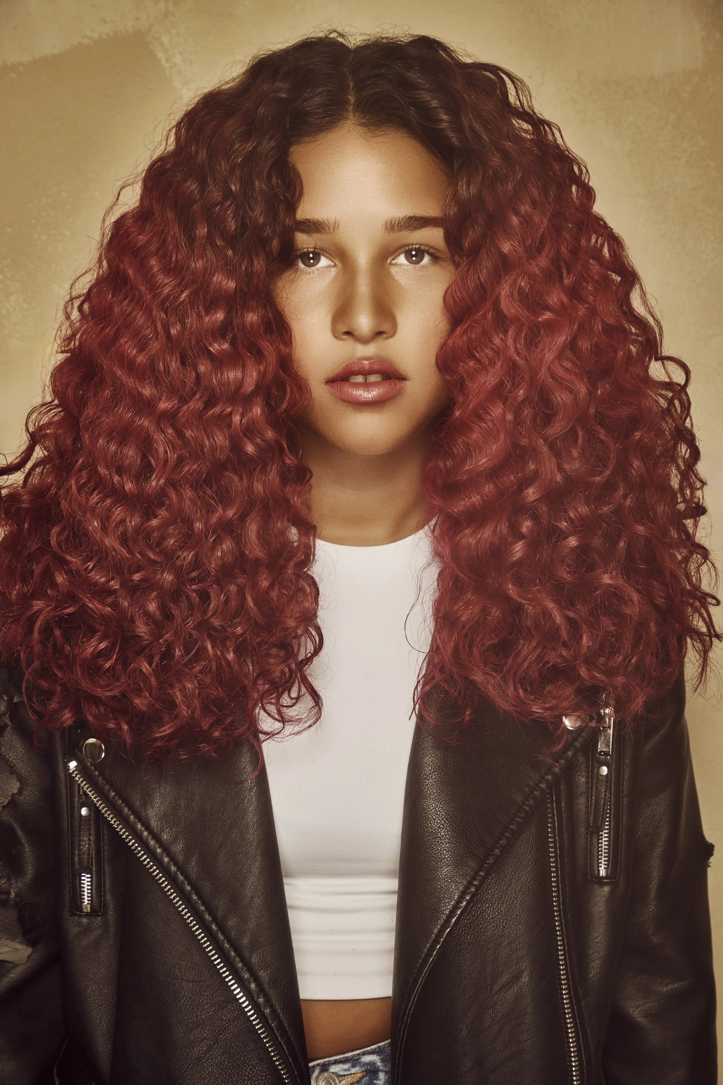 A woman with long, curly red hair, wearing a black leather jacket and a white shirt, looking into the camera.