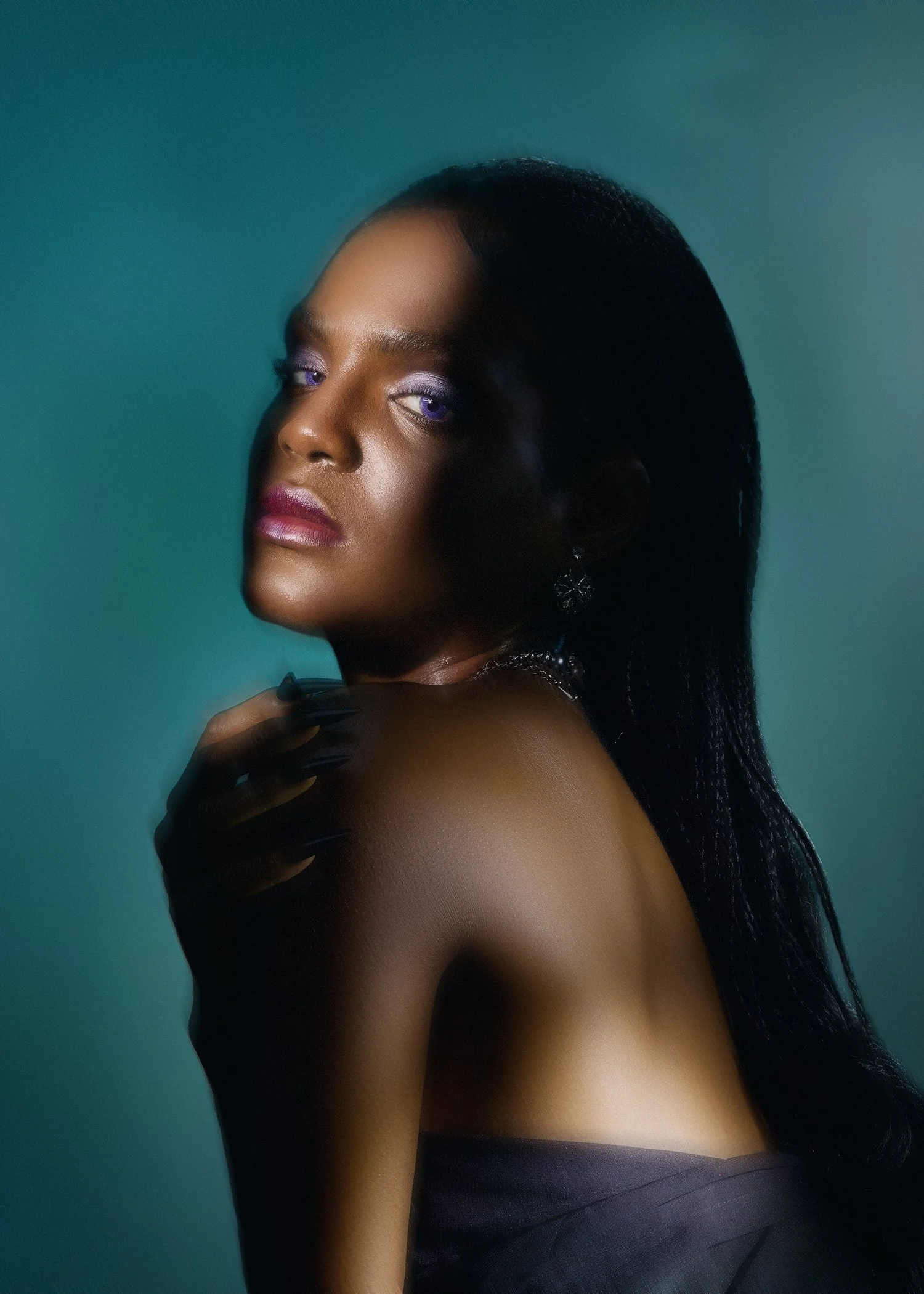 A portrait of a woman with long black hair, purple eyeshadow, and dark lipstick, posing against a gradient teal background.