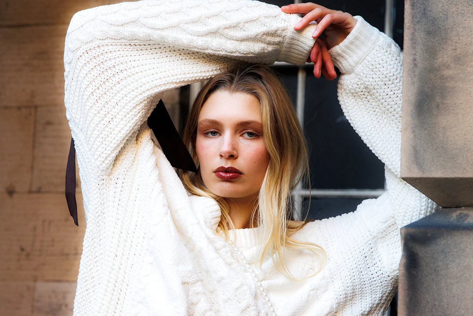 A young woman with blonde hair and blue eyes wearing a white knitted sweater, resting her arm on her head and leaning against a stone structure, with an urban background.