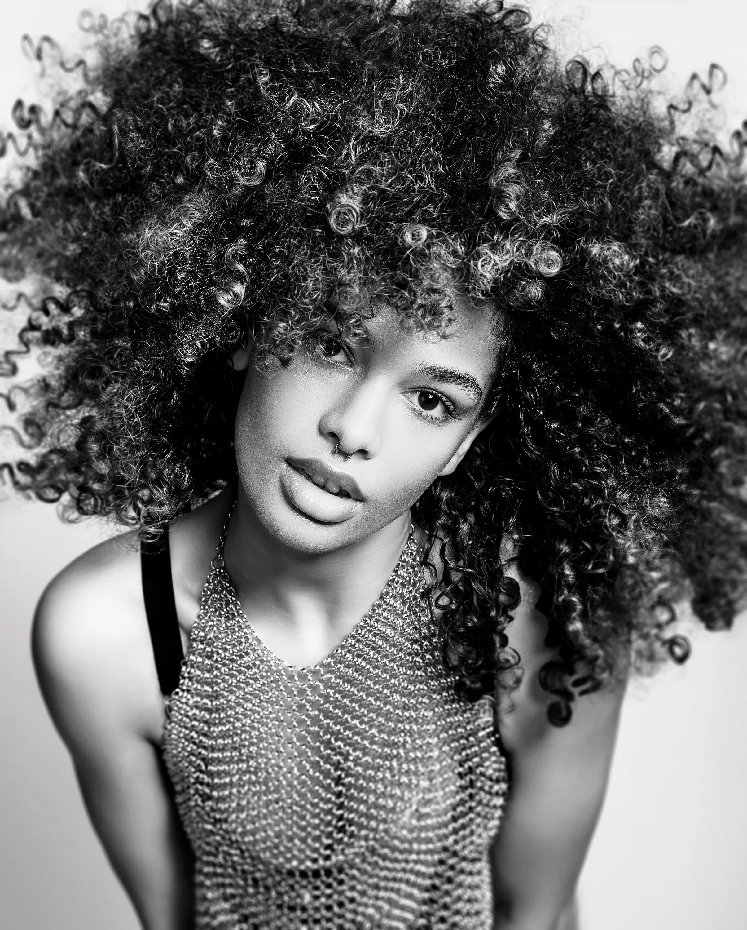 A woman with voluminous, curly hair, wearing a chainmail top and a septum ring, looking at the camera with a slightly open mouth.