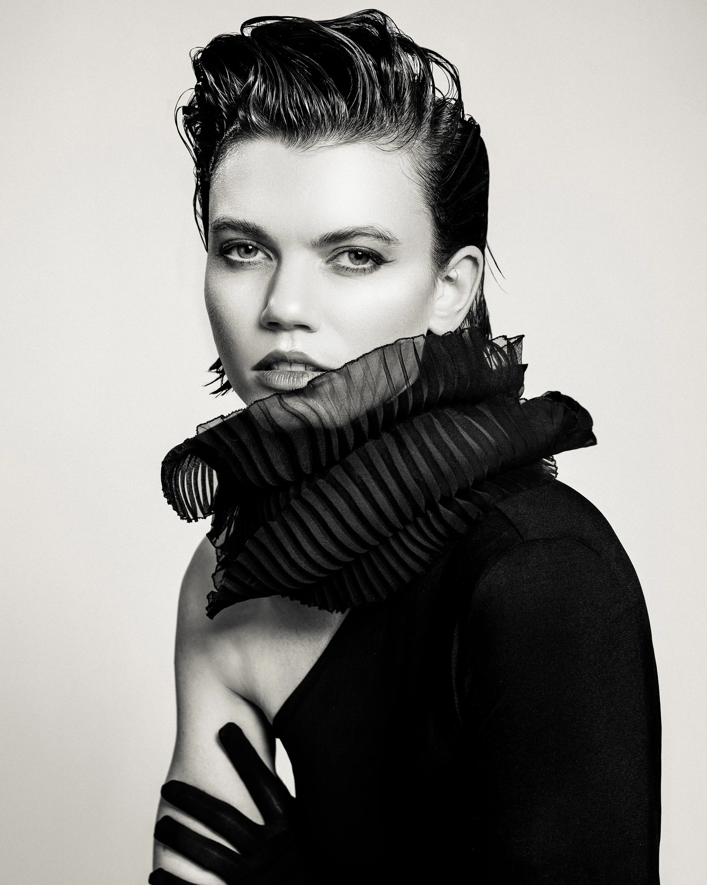 A black and white portrait of a woman with short, slicked-back hair, wearing a ruffled, striped black scarf around her neck and a black top.