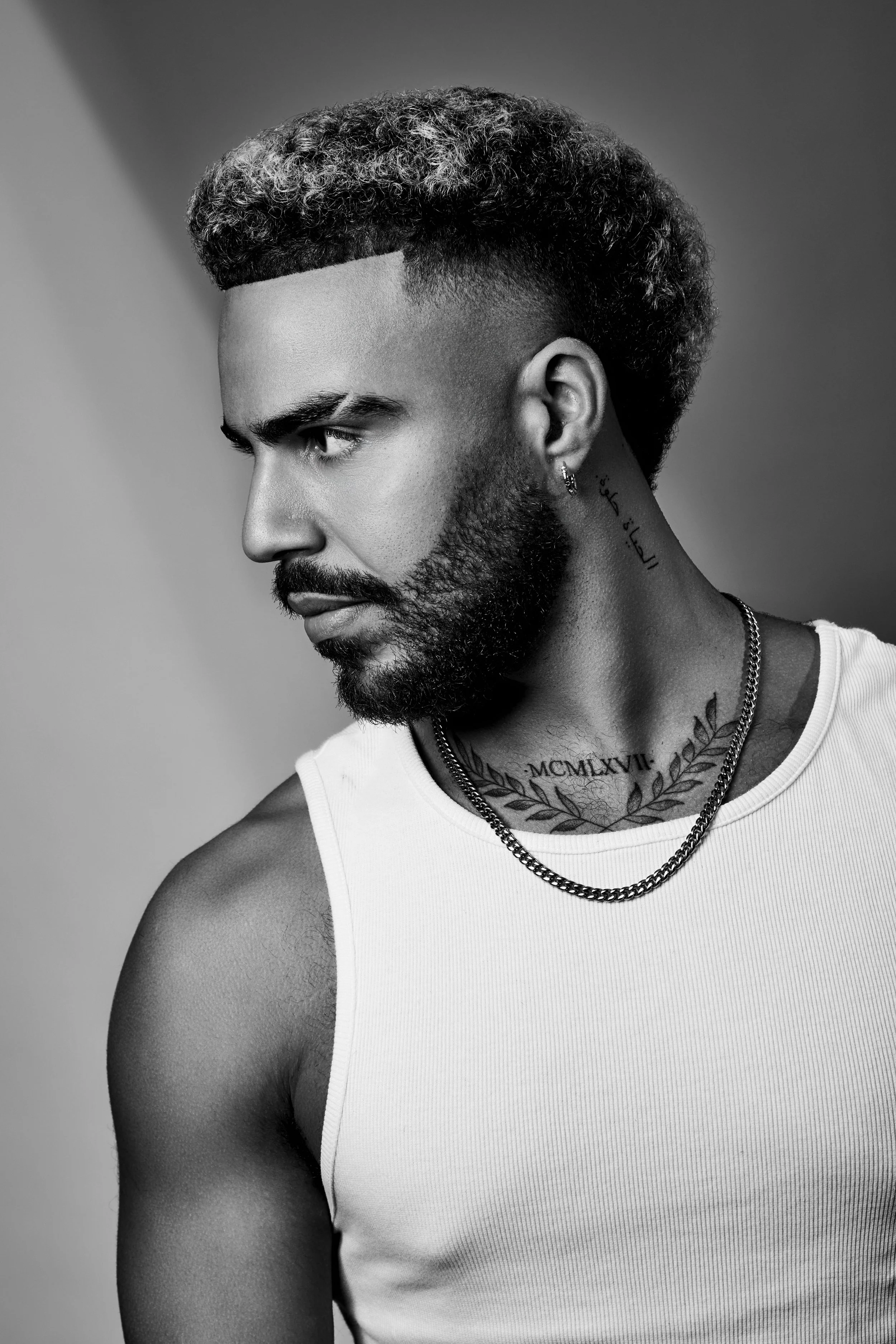 Black and white portrait of a man with a styled haircut, beard, and tattoos on his neck and chest, wearing a white tank top and chain necklace.