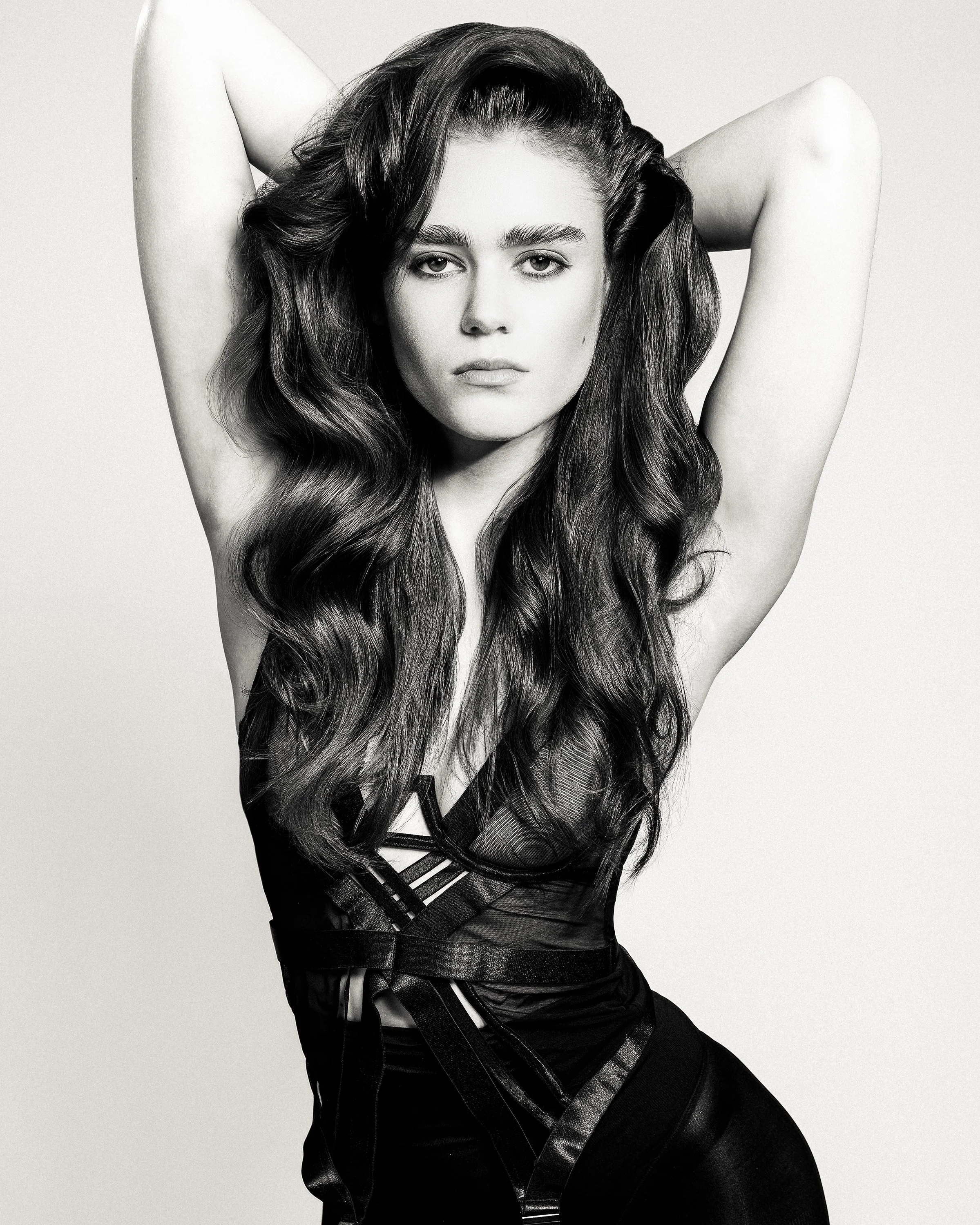 Black and white photo of a woman with long wavy hair, wearing a black sheer top, posing with arms behind her head.