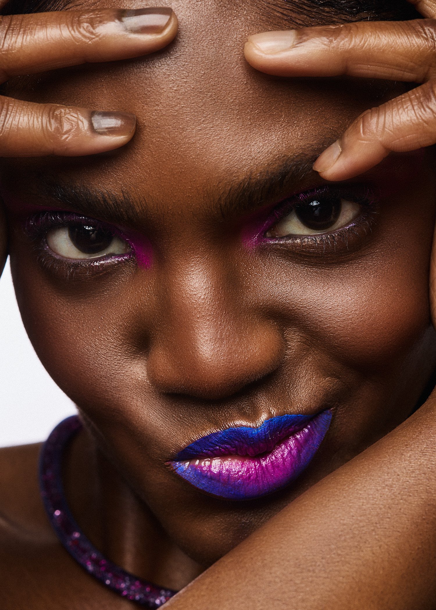 Close-up of a woman with dark skin, wearing colorful makeup including purple and blue lipstick and pink eyeshadow, with her hands framing her face.