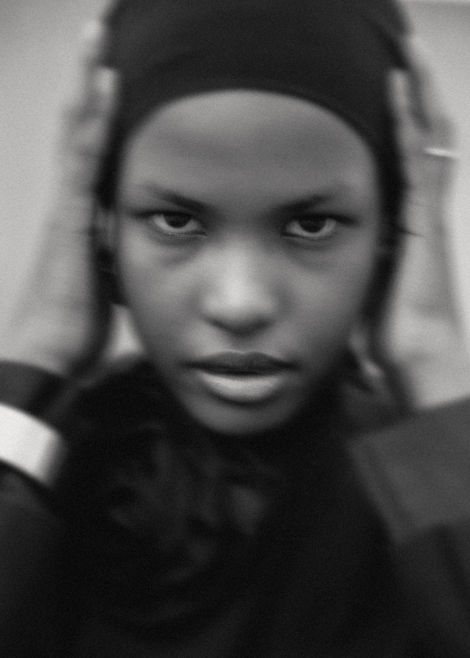 A close-up black and white portrait of a woman with intense eye contact, holding her head with both hands, with visible dark hair and a furrowed brow.