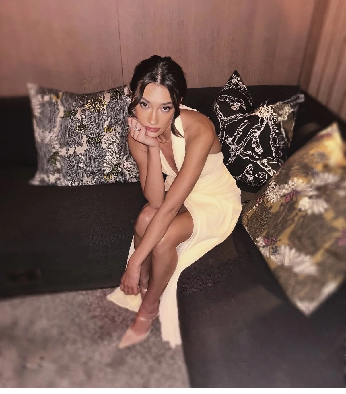 A woman in a cream-colored dress sitting on a black couch, resting her chin on her hand, surrounded by decorative pillows with intricate patterns.