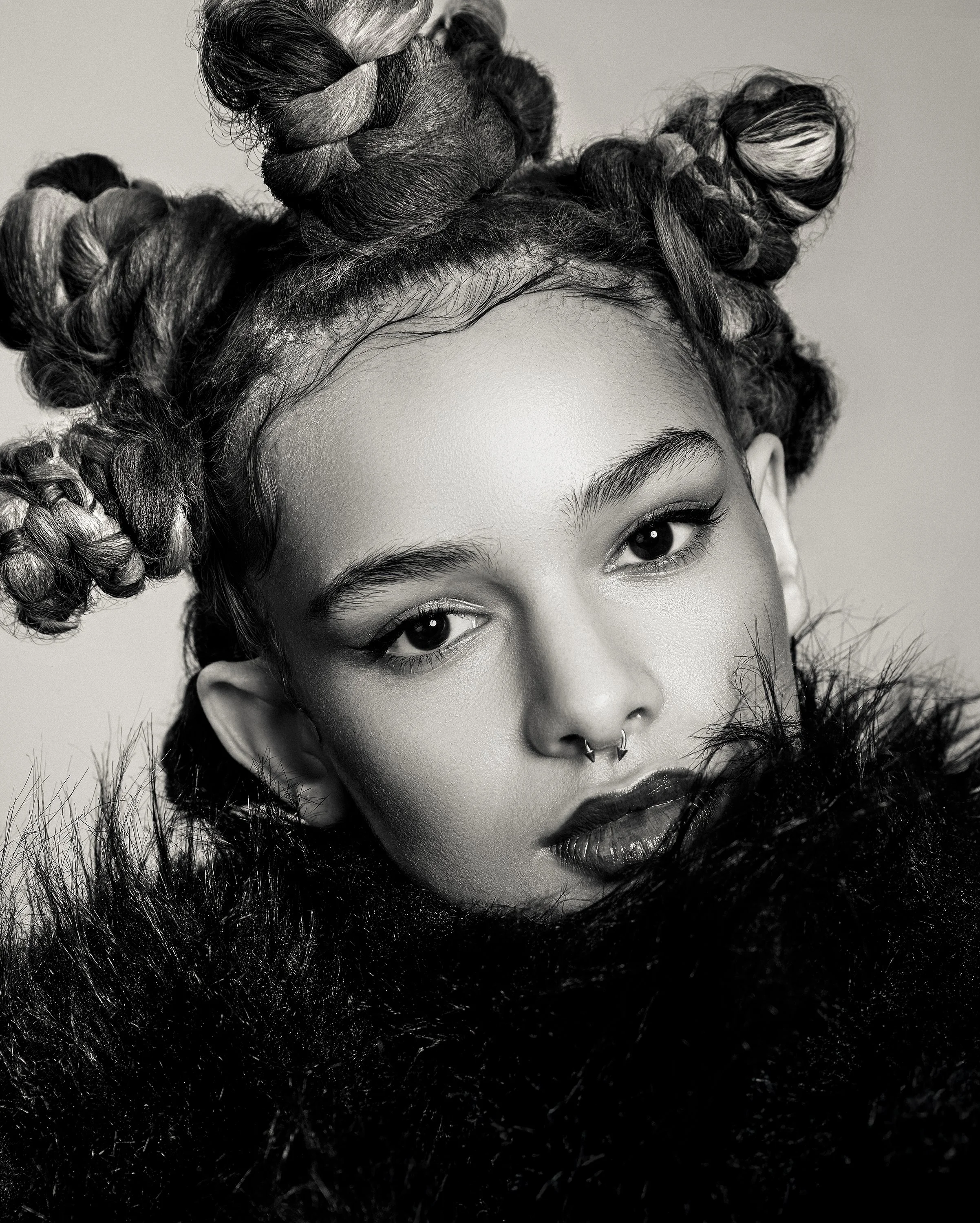 Black and white portrait of a young woman with curly hair styled in buns, wearing dark lipstick, a septum piercing, and a fur garment.