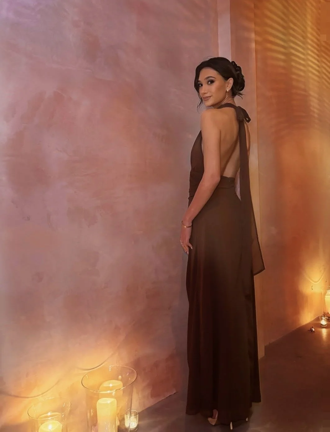 A woman in a brown halter top and matching long skirt standing indoors near a textured pink and orange wall, with candles on the floor.