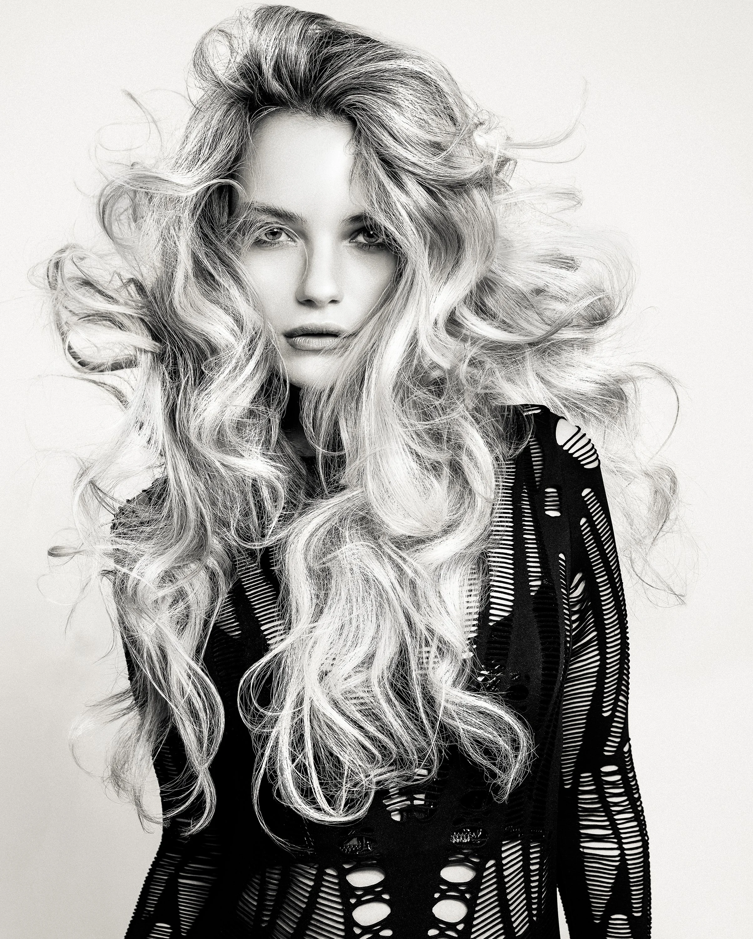 A black-and-white portrait of a woman with long, wavy hair wearing a black, ripped, semi-transparent top.