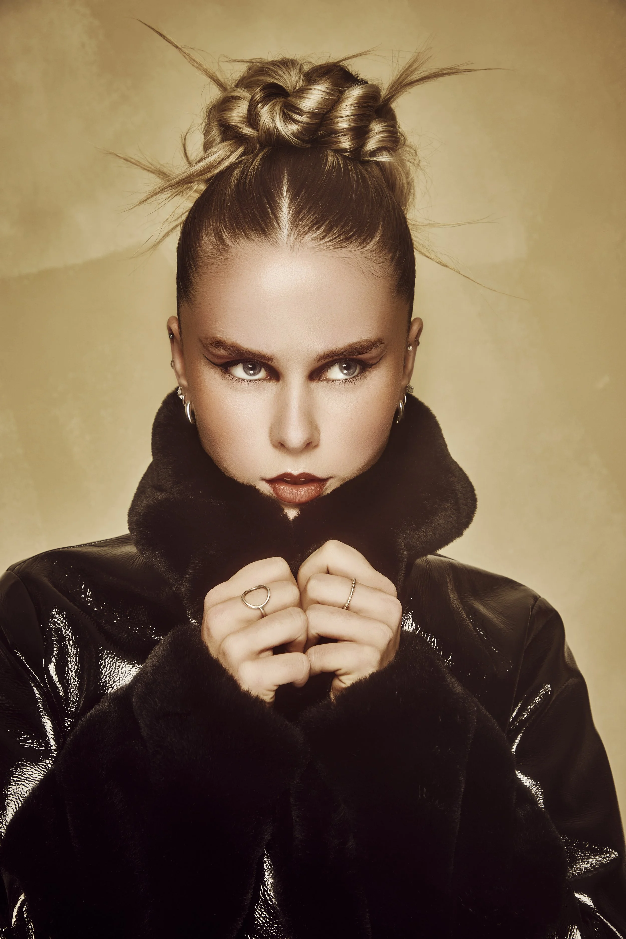 A woman with styled hair, intense eye makeup, and red lipstick wearing a black fur-collared jacket, holding the collar with her hands.