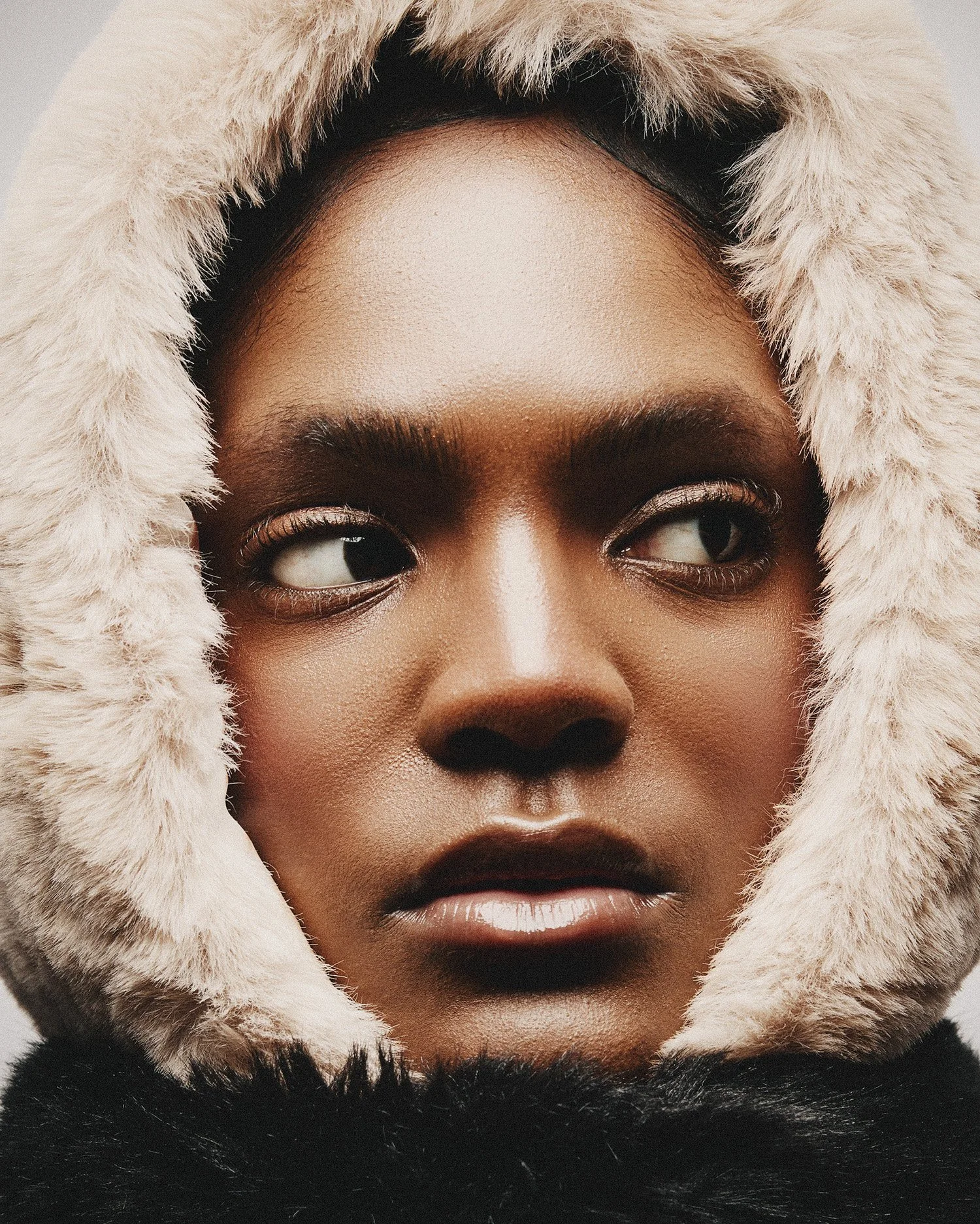 Close-up of a woman's face with makeup, wearing a fur-lined hood, looking to the side.