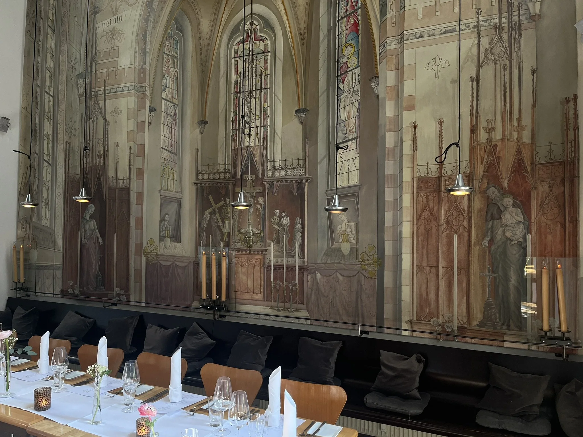 Eleganter Speisesaal mit historischer Wandmalerei im gotischen Stil, geschmückt mit Kerzen und moderner Beleuchtung. Der Tisch ist stilvoll mit Gläsern, Besteck und weißen Servietten gedeckt, ergänzt durch kleine Blumenarrangements.