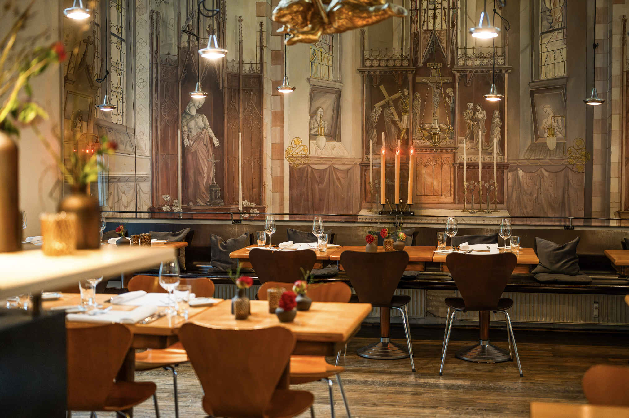Innenansicht eines stilvollen Restaurants mit Holztischen, Stühlen, gedeckten Tischen und Kerzenständern, an einer Wand mit aufwändigem Wandgemälde im gotischen Stil dekoriert.