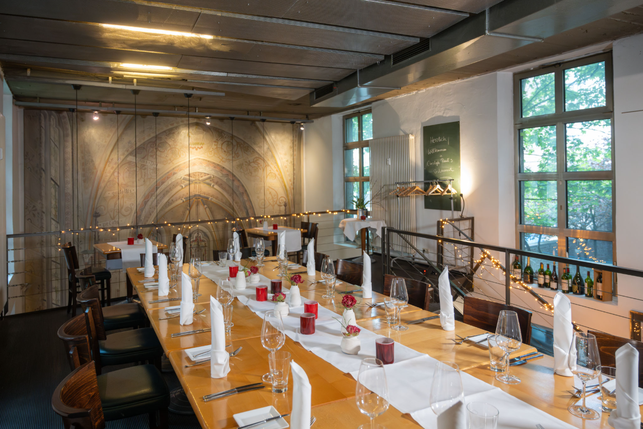 Eleganter Speisesaal mit langem, gedecktem Tisch, dekoriert mit roten Kerzen und Servietten. Im Hintergrund sind dekorative Wände und große Fenster zu sehen. An der Wand hängen Kleiderbügel und ein Menübrett.