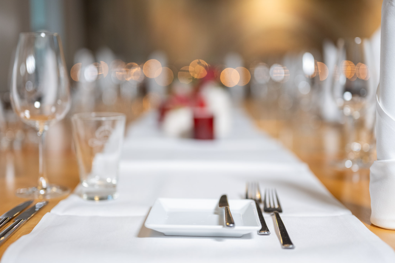 Ein eleganter, festlich gedeckter Tisch mit einem Teller, Besteck, einem Glas und einer Serviette. Im Hintergrund sind weitere Gläser und unscharfe Lichter zu sehen.