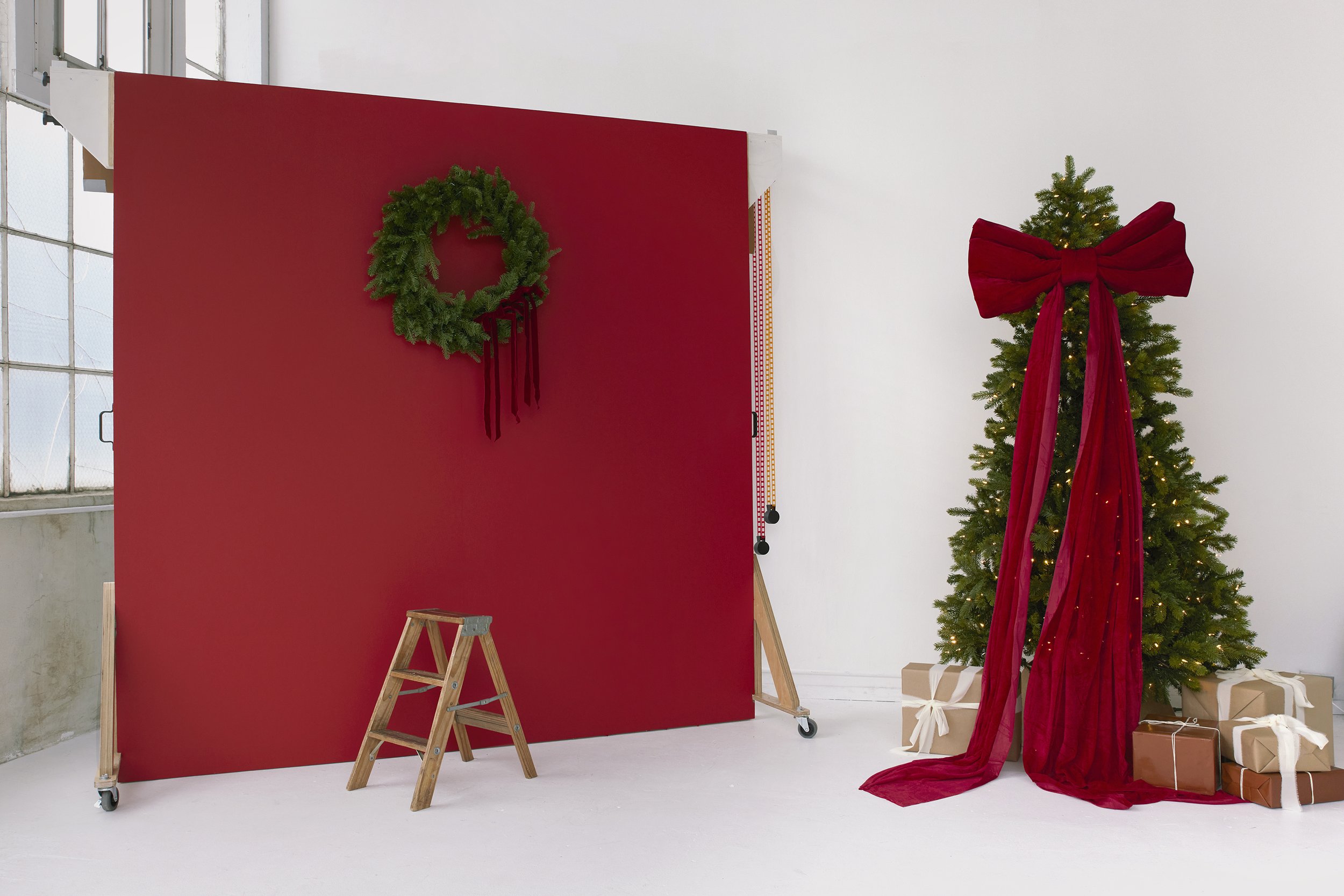 Christmas scene with a decorated tree draped in a large red bow, presents underneath, and a red backdrop with a green wreath hanging.