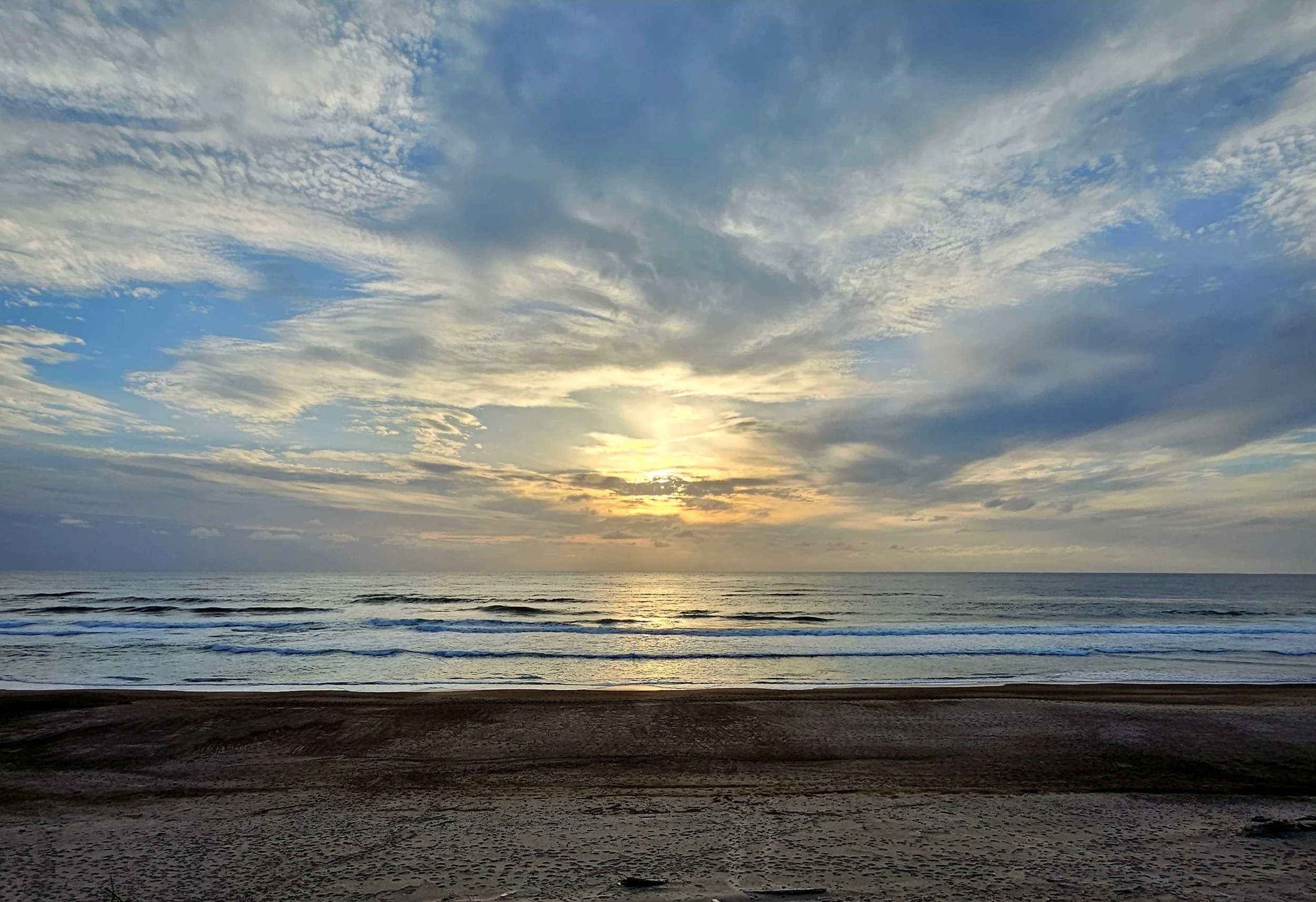 A beach scene at sunrise with the sun partially hidden behind clouds, calm ocean waves, and a sandy shore in the foreground.