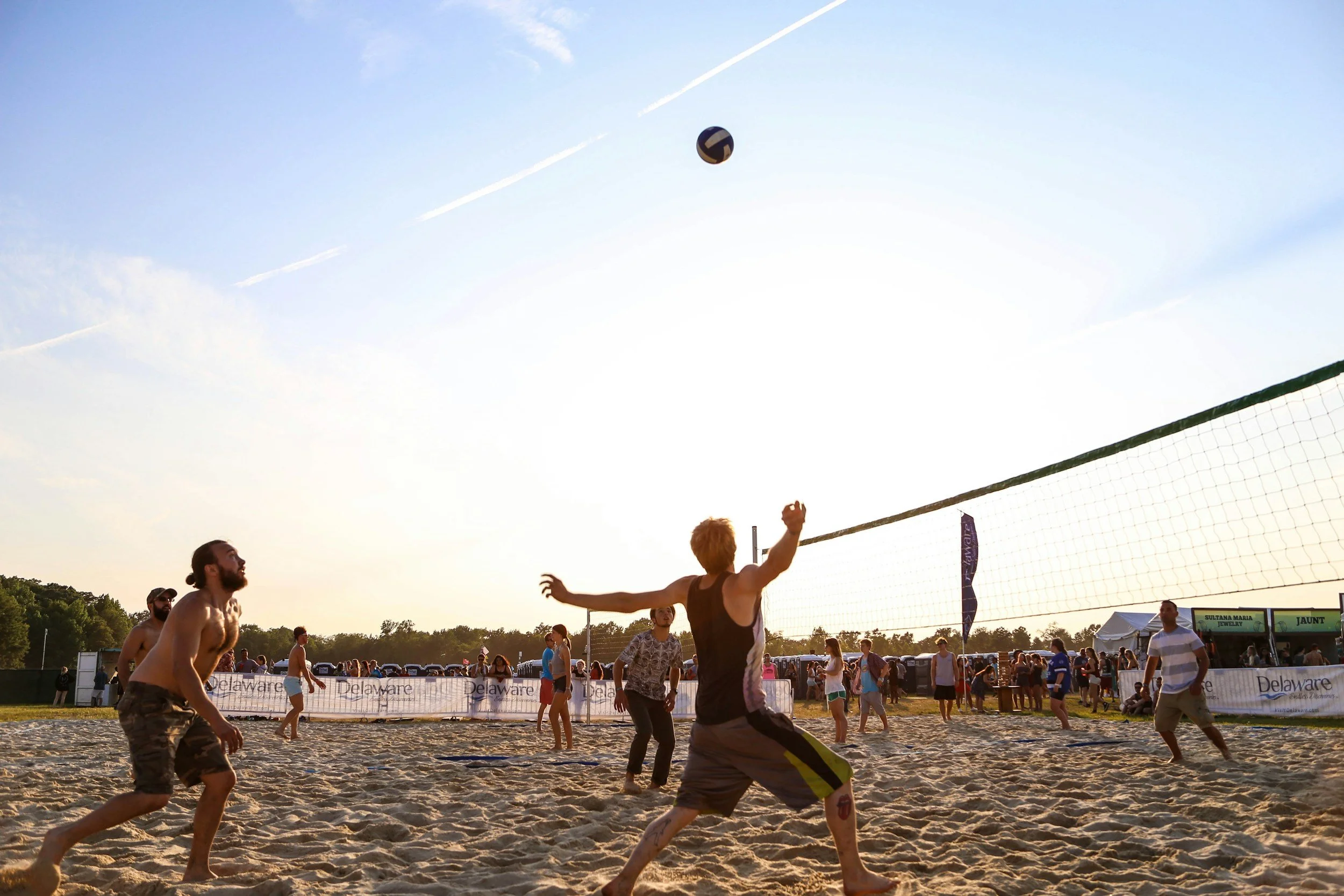 People playing beach volleyball under a clear sky, with a net and a crowd watching in the background.
