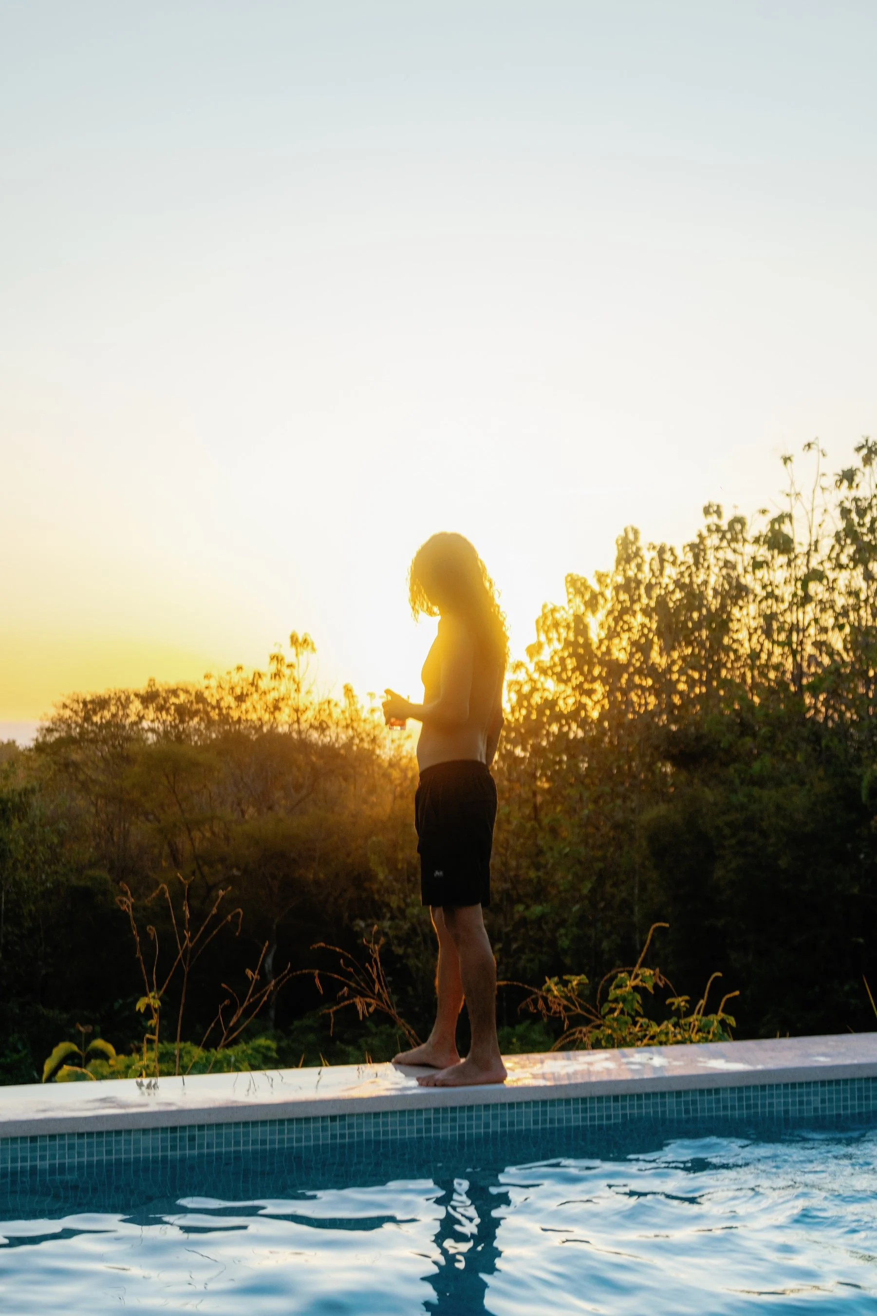 A person with long hair standing on the edge of a swimming pool during sunset, looking at their phone.