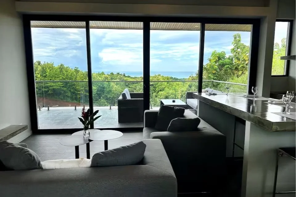 Living room with large glass sliding doors opening to a balcony with a view of green trees and the ocean in the distance.