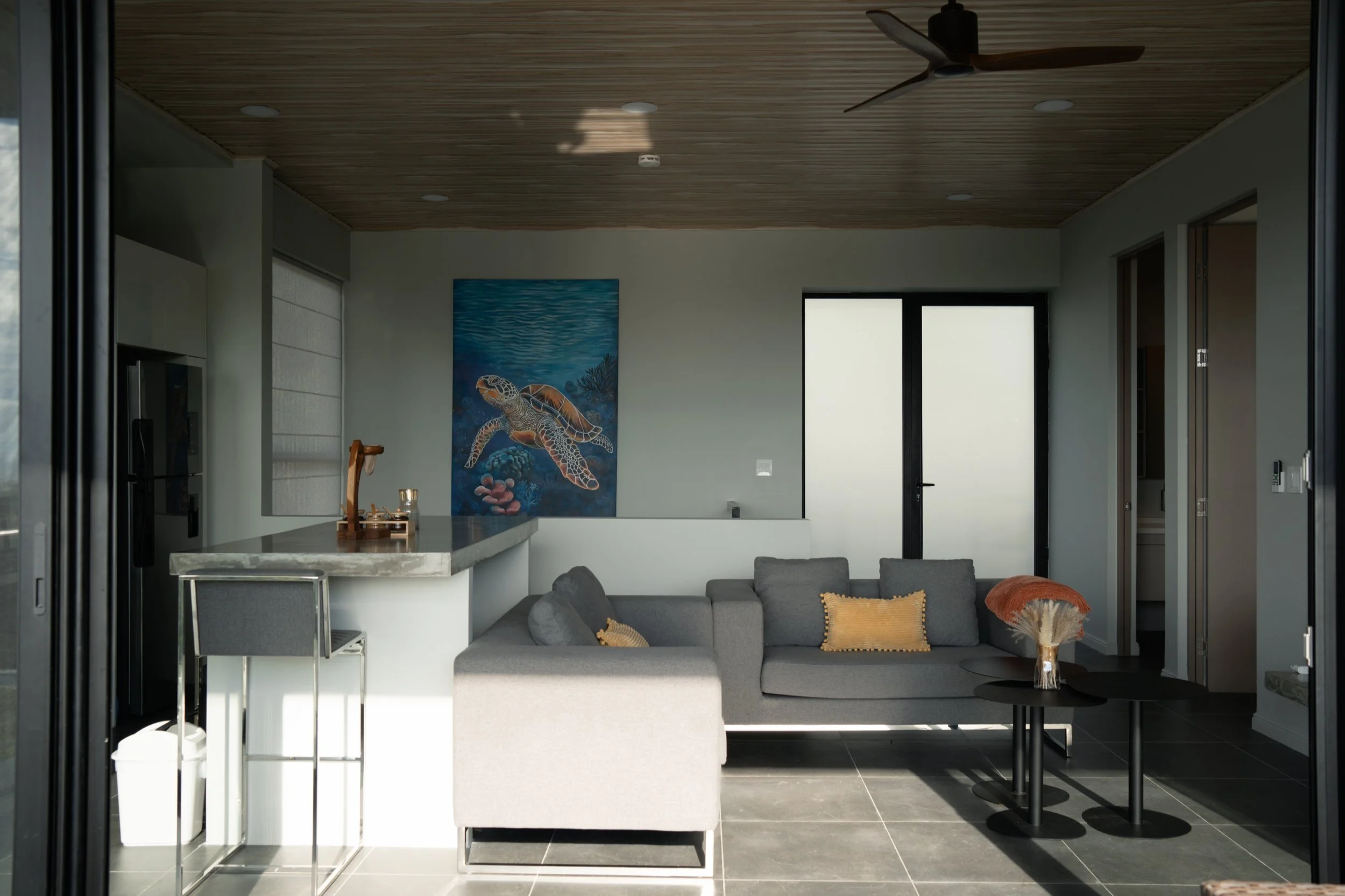 Modern living room with gray sofa, three throw pillows, black coffee tables, tropical painting of a turtle on the wall, large glass door, wooden ceiling, ceiling fan, and decorative vase with dried flowers.