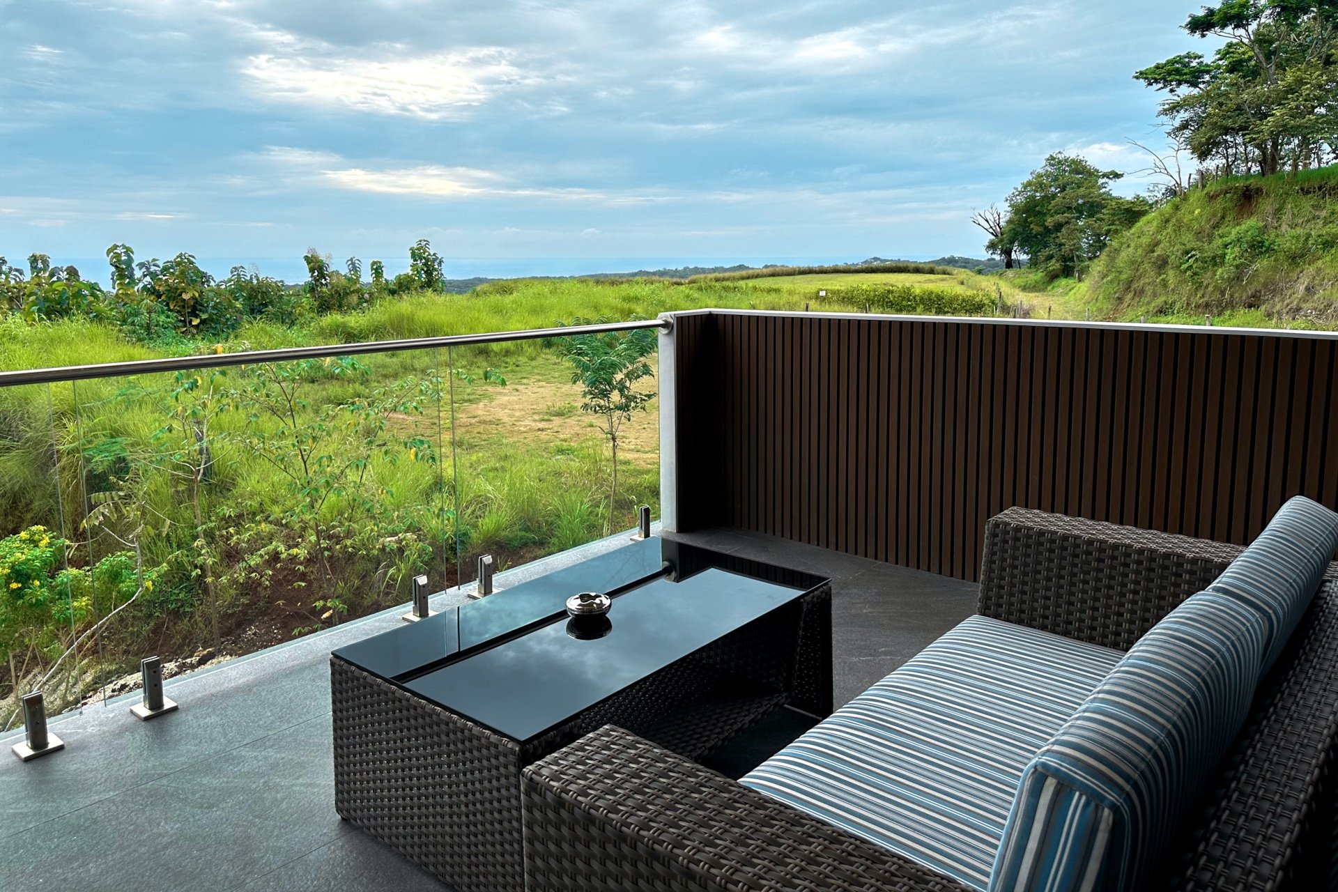 Balcony with wicker sofa with striped cushion, glass-top table with ashtray, overlooking green fields and trees under cloudy sky.
