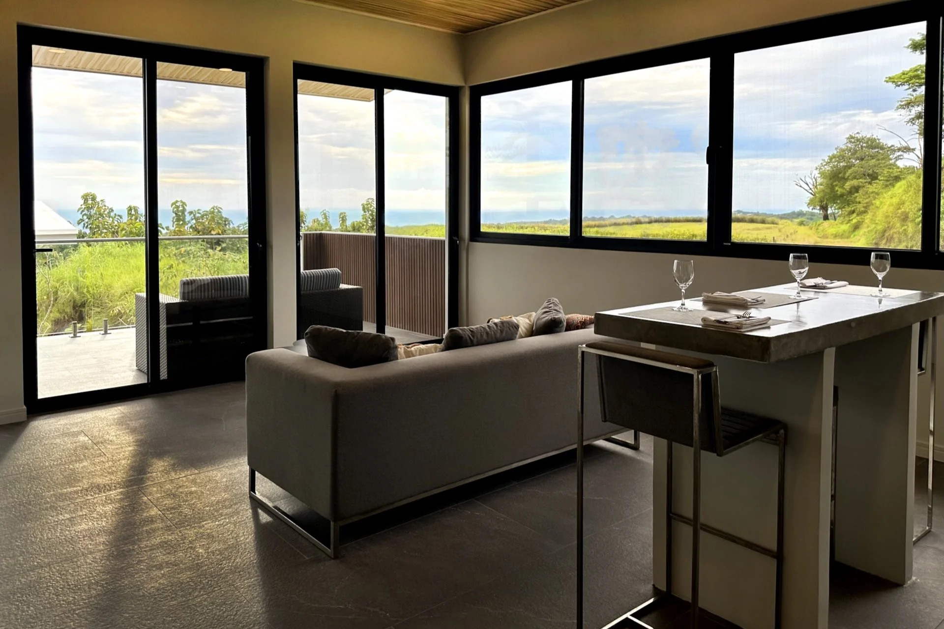 Living room with large windows overlooking a green landscape, a white sofa with pillows, a dining counter set with wine glasses and utensils, a balcony with outdoor seating.