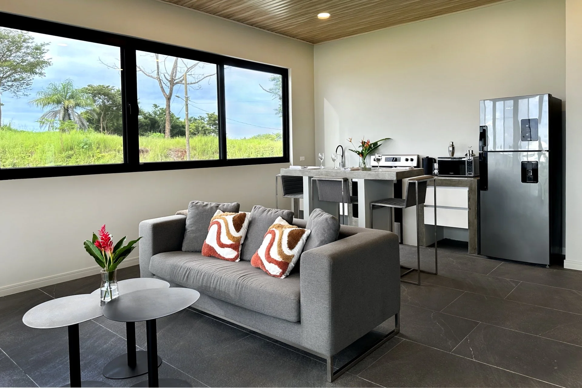 Modern apartment living room with large window, grey sofa with patterned cushions, small round coffee table with a flower arrangement, and an open kitchen with a refrigerator, microwave, and bar stools.