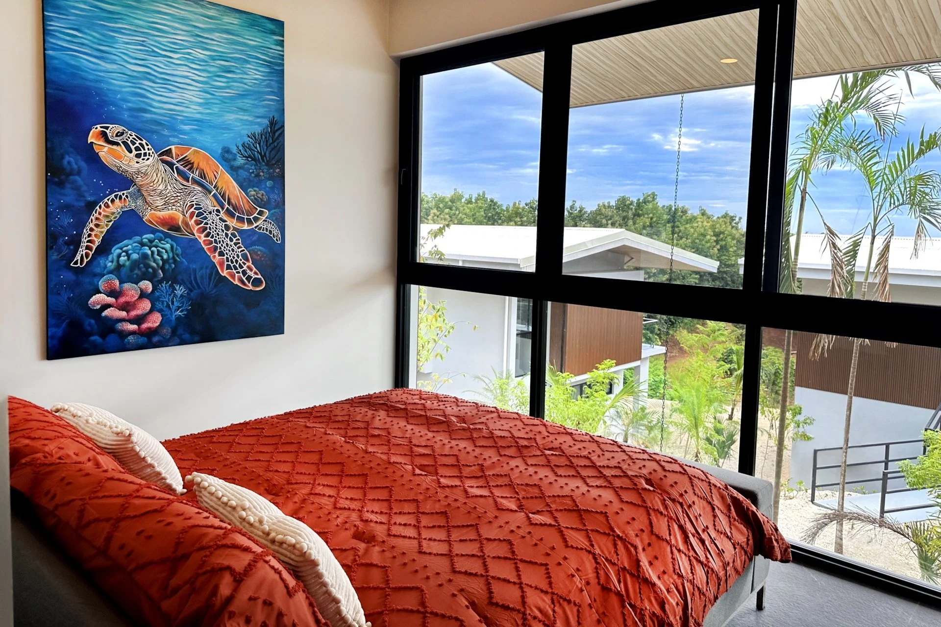 A bedroom with a bed featuring an orange quilt and white pillows, a colorful painting of a sea turtle on the wall, and large windows with a view of trees and modern buildings outside.