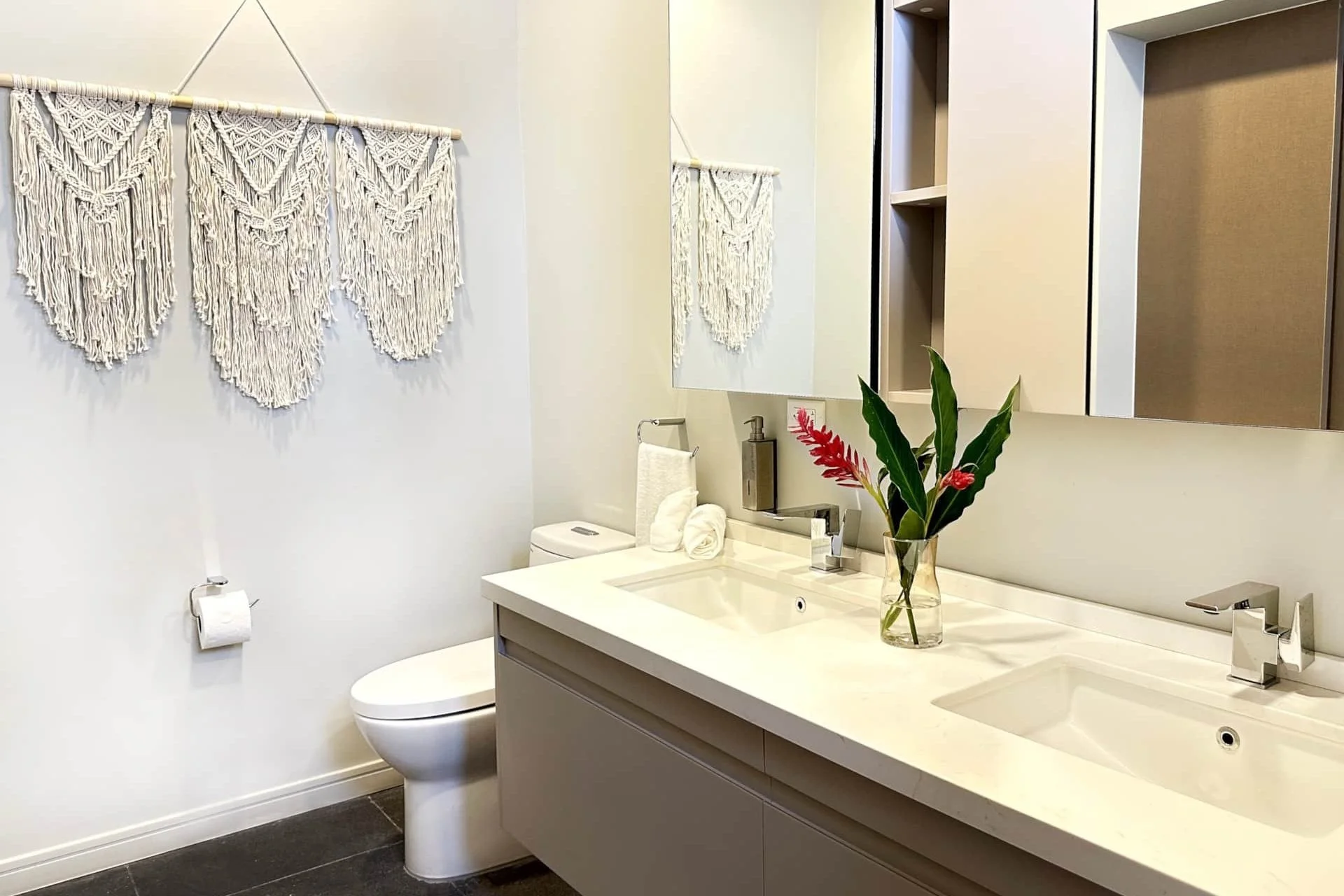 Modern bathroom with white walls, double sink vanity, large mirror, decorative flower vase, toilet, and macramé wall art.