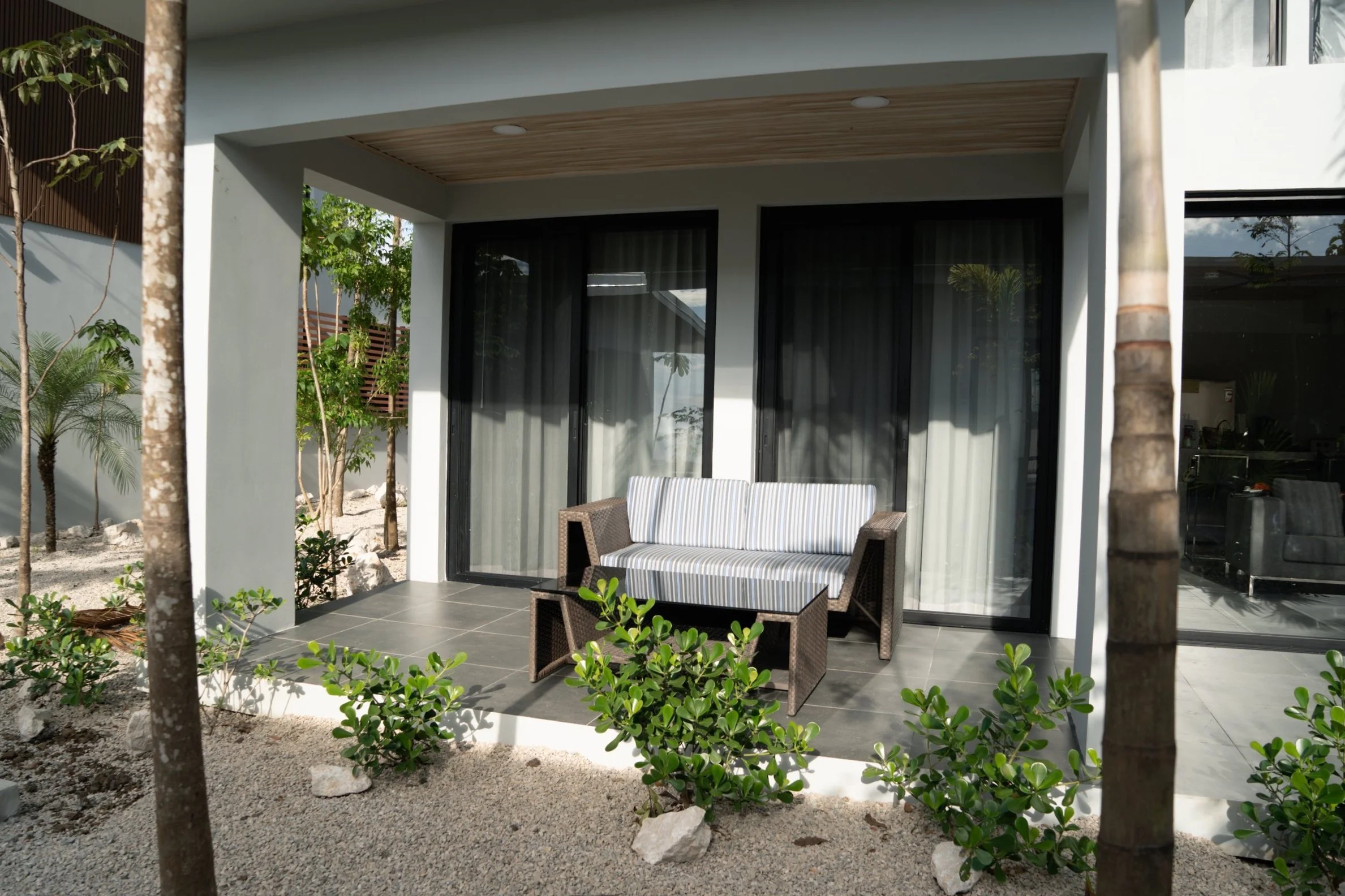 Modern patio with a striped armchair, matching loveseat, and coffee table, surrounded by greenery and trees, with sliding glass doors leading inside.