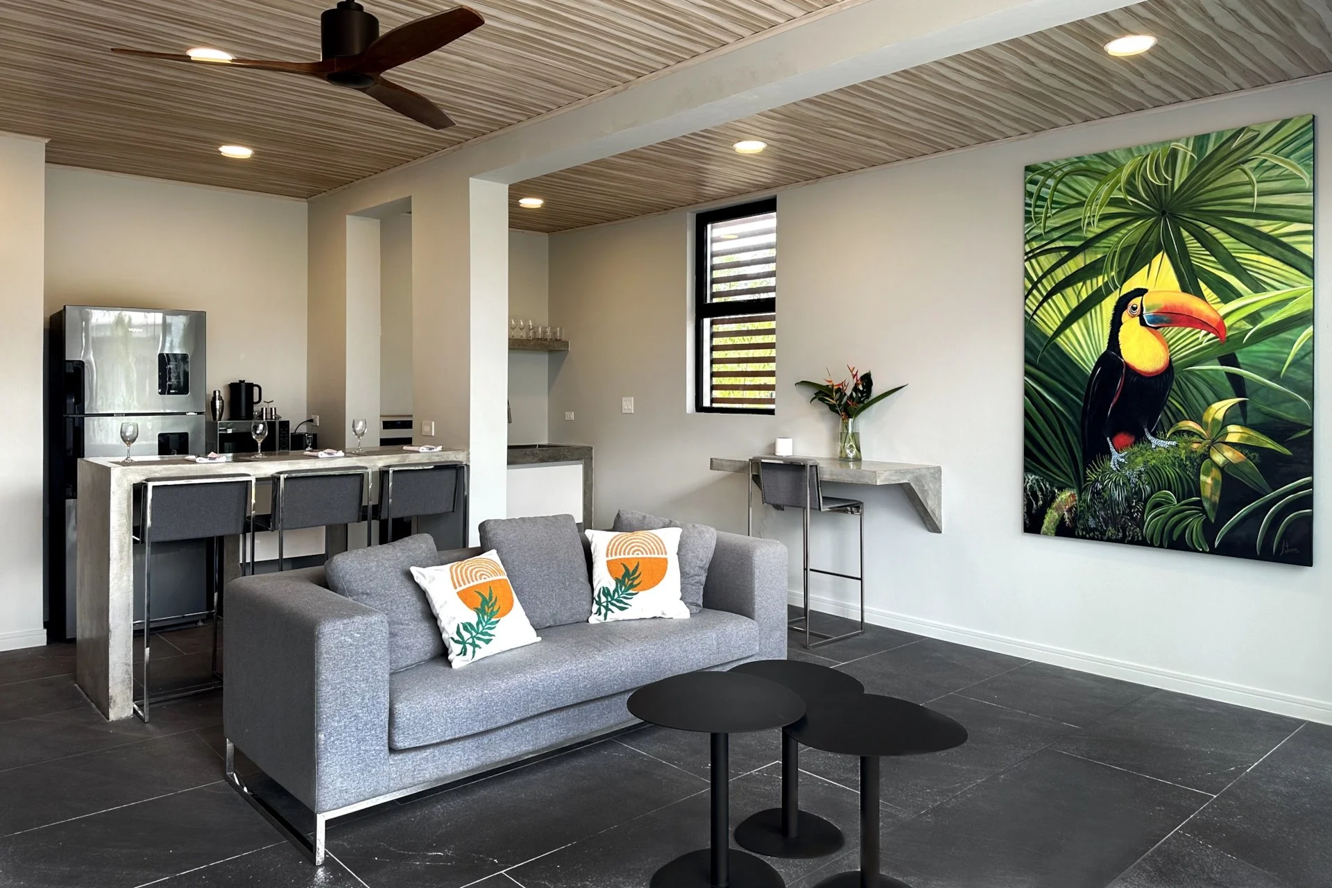 Modern living room with gray sofa, black coffee tables, a large colorful toucan painting on the wall, a small desk with a flower vase, and a kitchen area with high chairs, refrigerator, and window.