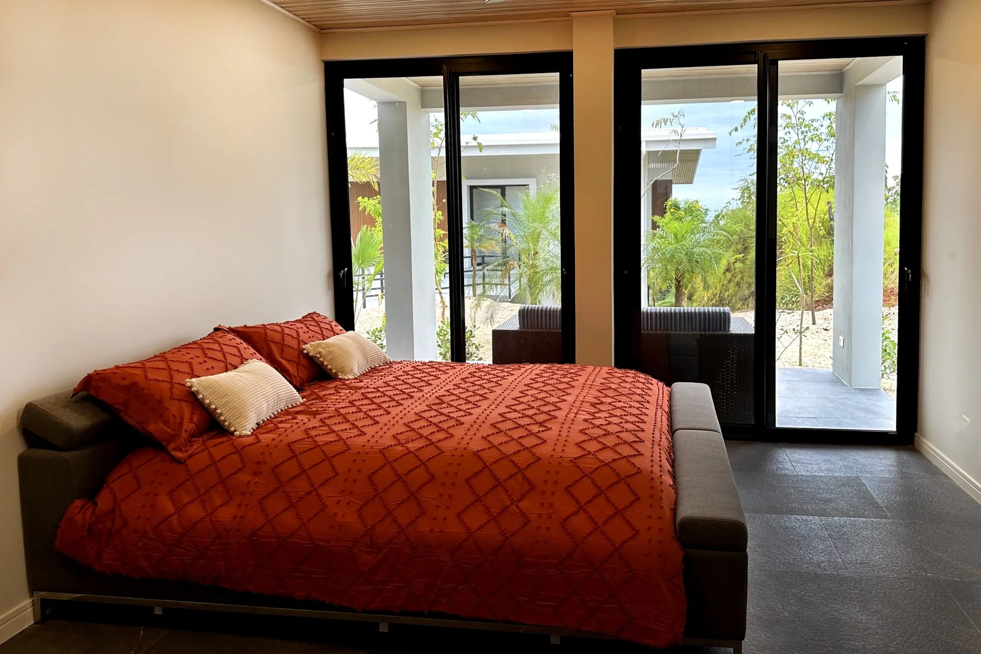 A bedroom with a large bed covered with an orange quilt and white pillows, positioned near glass sliding doors that open to a patio with outdoor seating and plants, and a view of outside greenery.