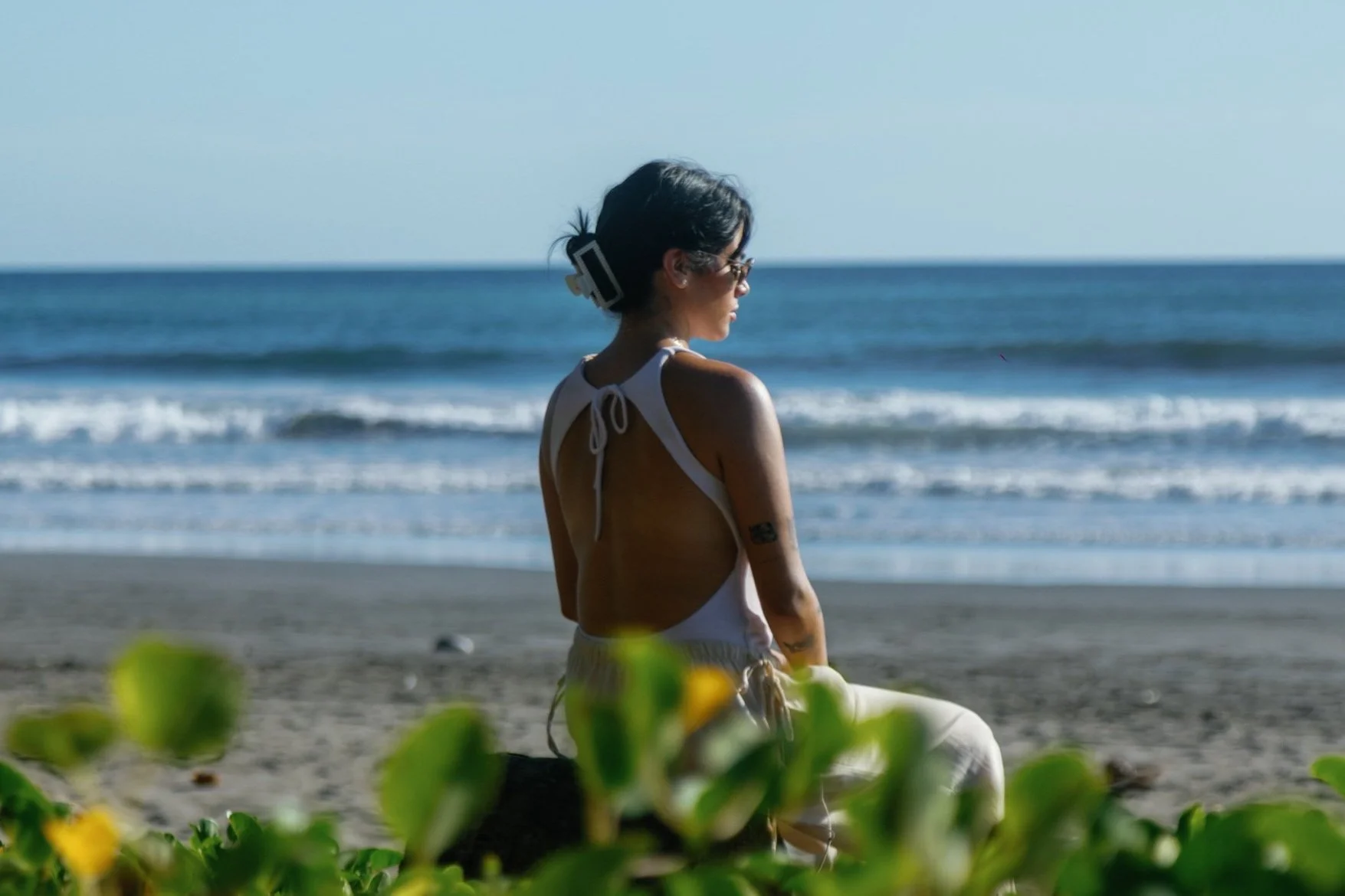 Woman sitting on the beach near green plants, facing the ocean with waves, wearing sunglasses, a sleeveless top, and light-colored pants.