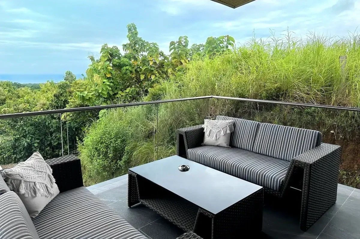 Outdoor patio with black wicker furniture including a sofa and a chair with striped cushions, a black coffee table, and a glass railing overlooking greenery and a distant view of the sky.