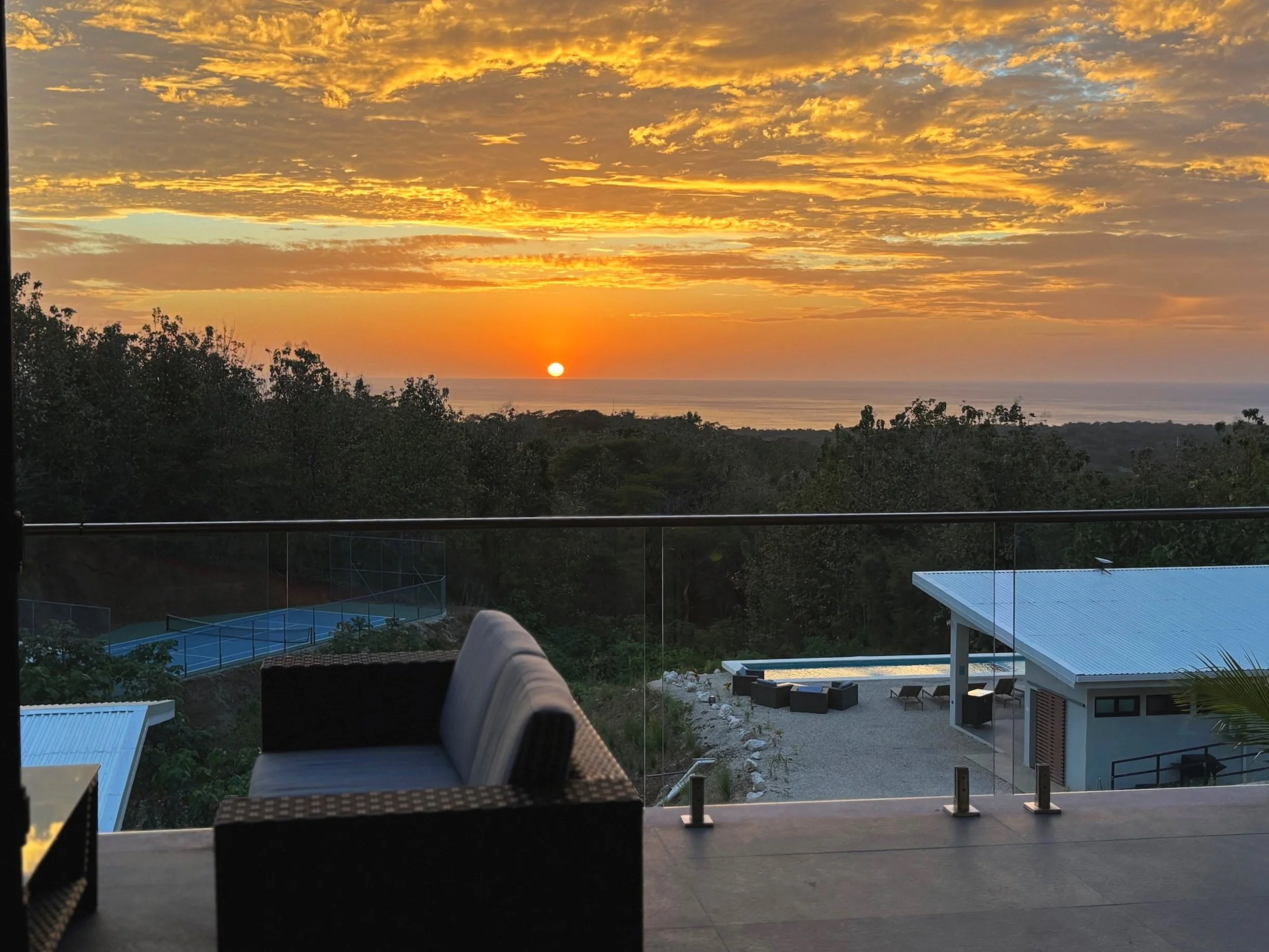 Sunset view over a treetops and ocean, with a glass balcony and outdoor furniture, including a cushioned chair and a table, in a modern house.