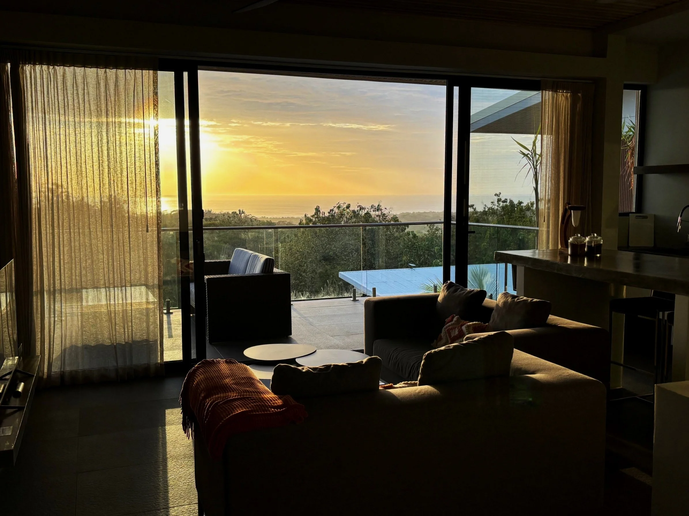 Living room with large sliding glass door opening to a balcony and view of sunset over a lush, green landscape